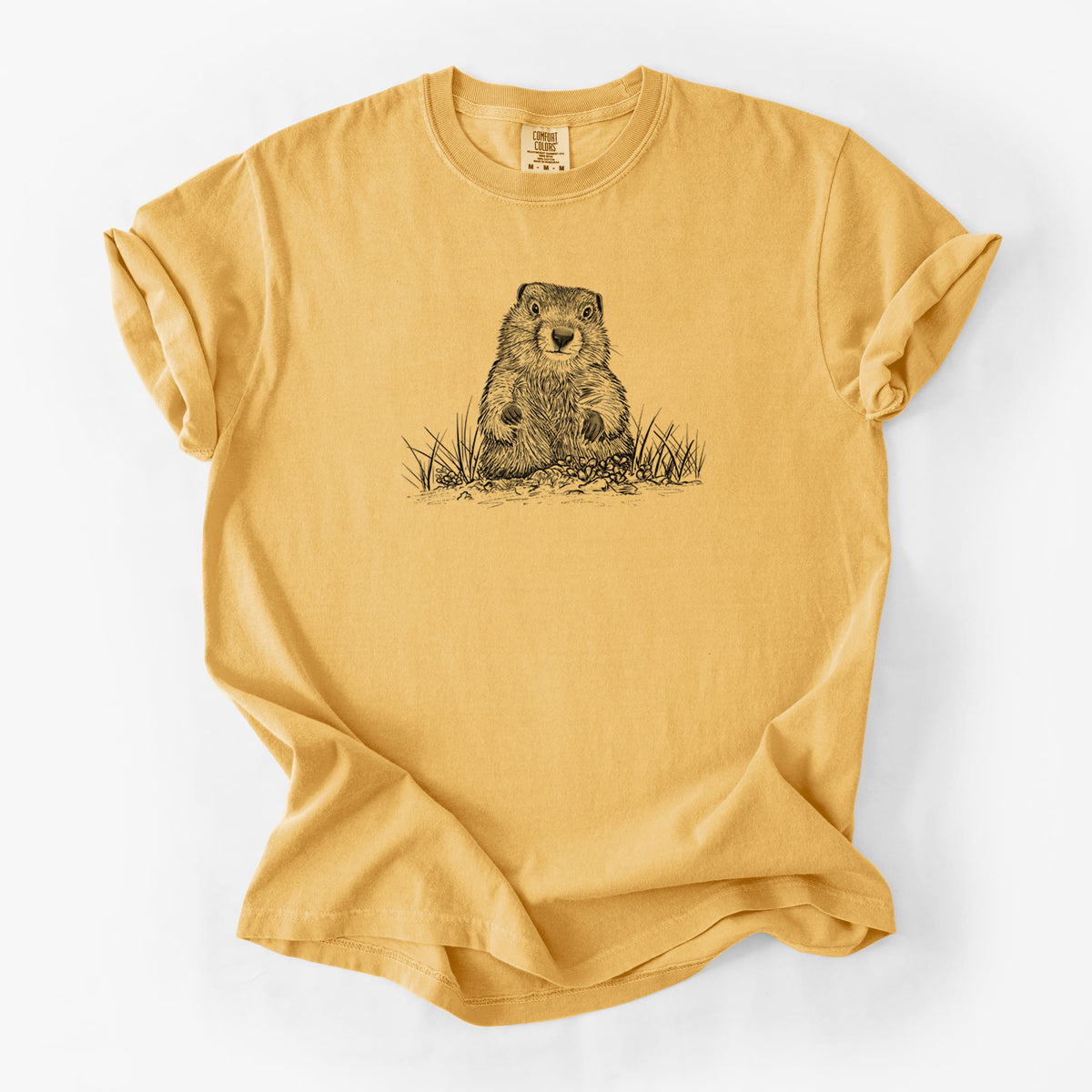 Golden wheat yellow Comfort Colors t-shirt laid flat, printed with a black ink illustration of a groundhog peeking out of its burrow among grass.