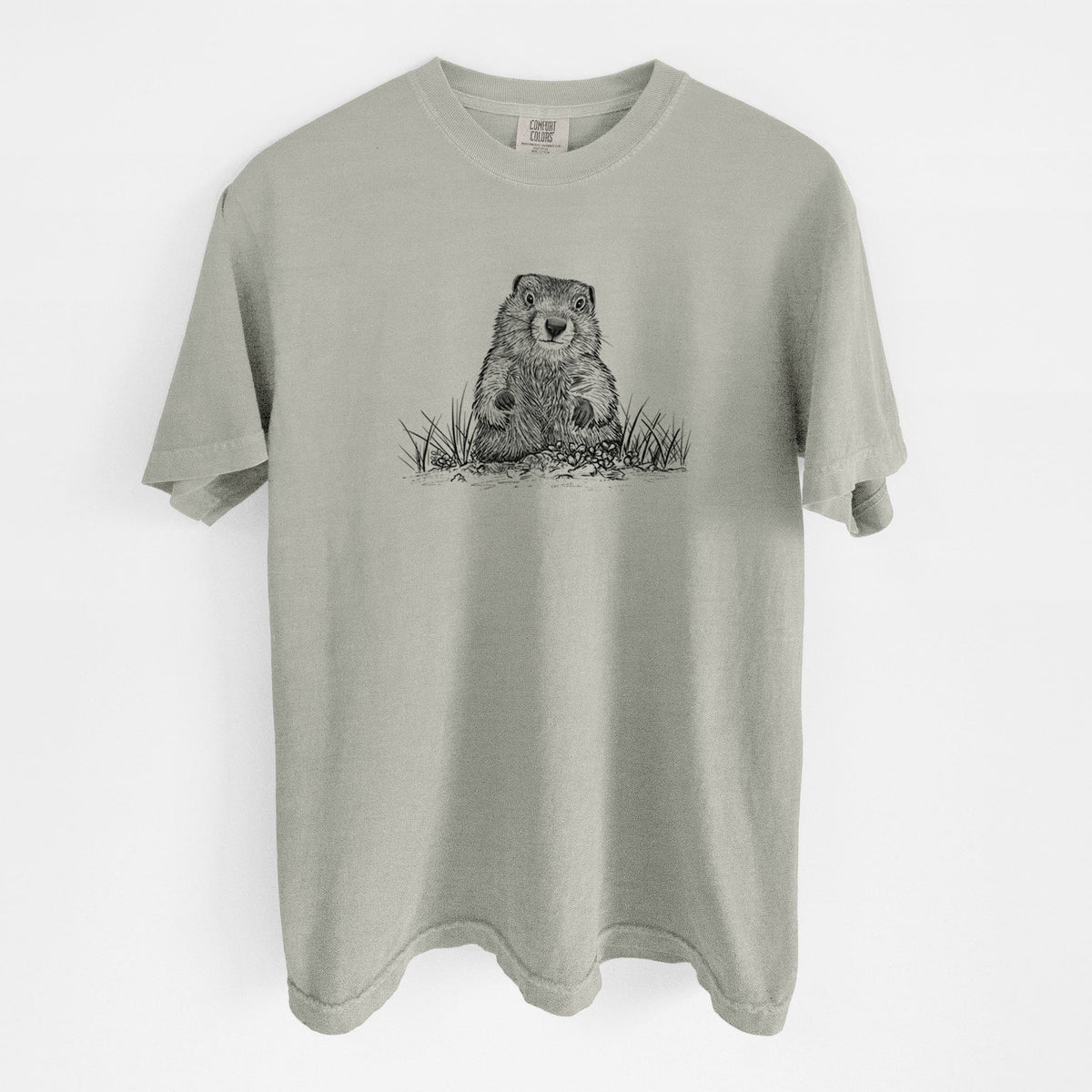 Sandstone beige Comfort Colors t-shirt laid flat, printed with a black ink illustration of a groundhog peeking out of its burrow among grass.