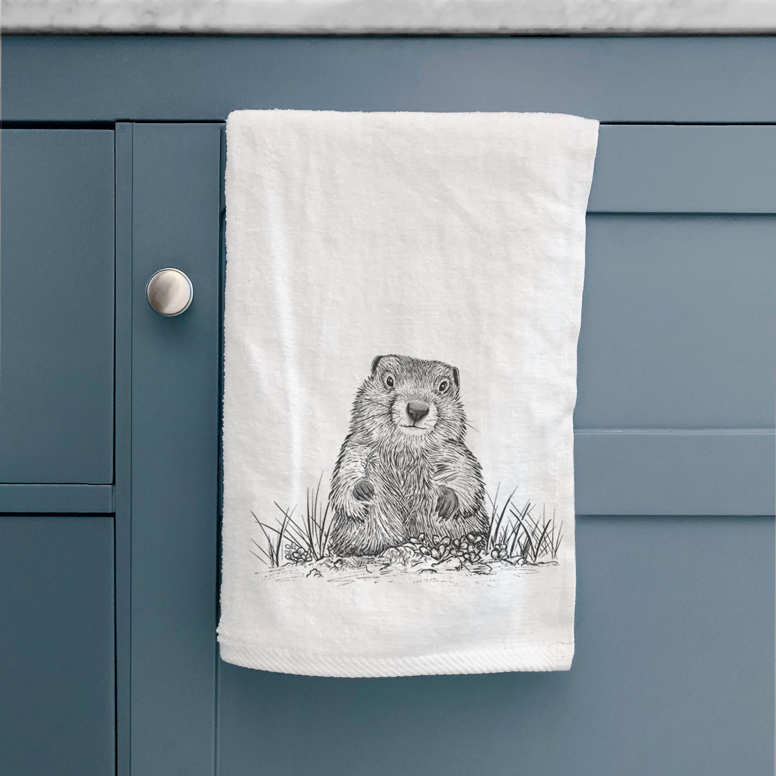 White hand towel printed with a black ink illustration of a groundhog peeking out of its burrow among grass.