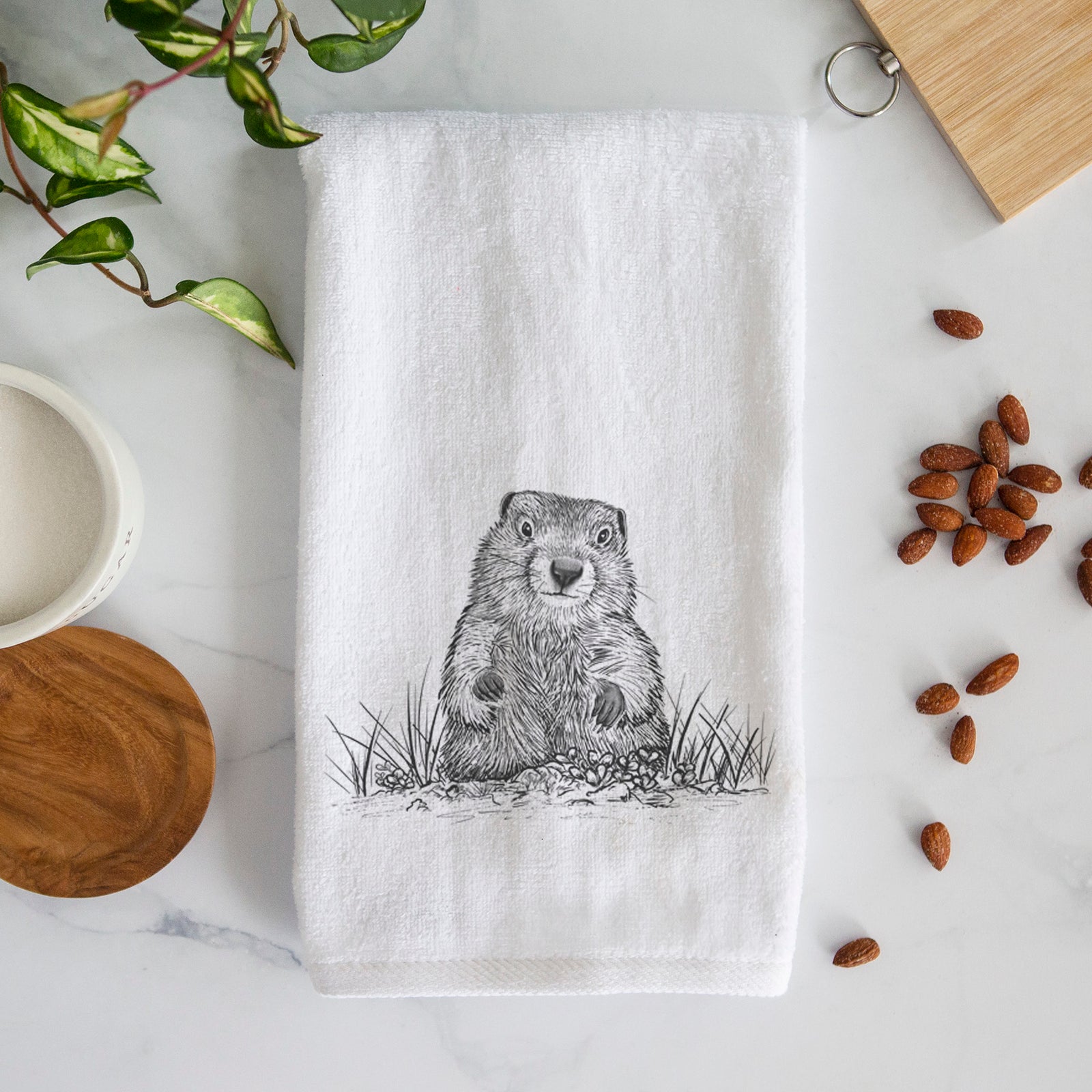 White hand towel printed with a black ink illustration of a groundhog peeking out of its burrow among grass.