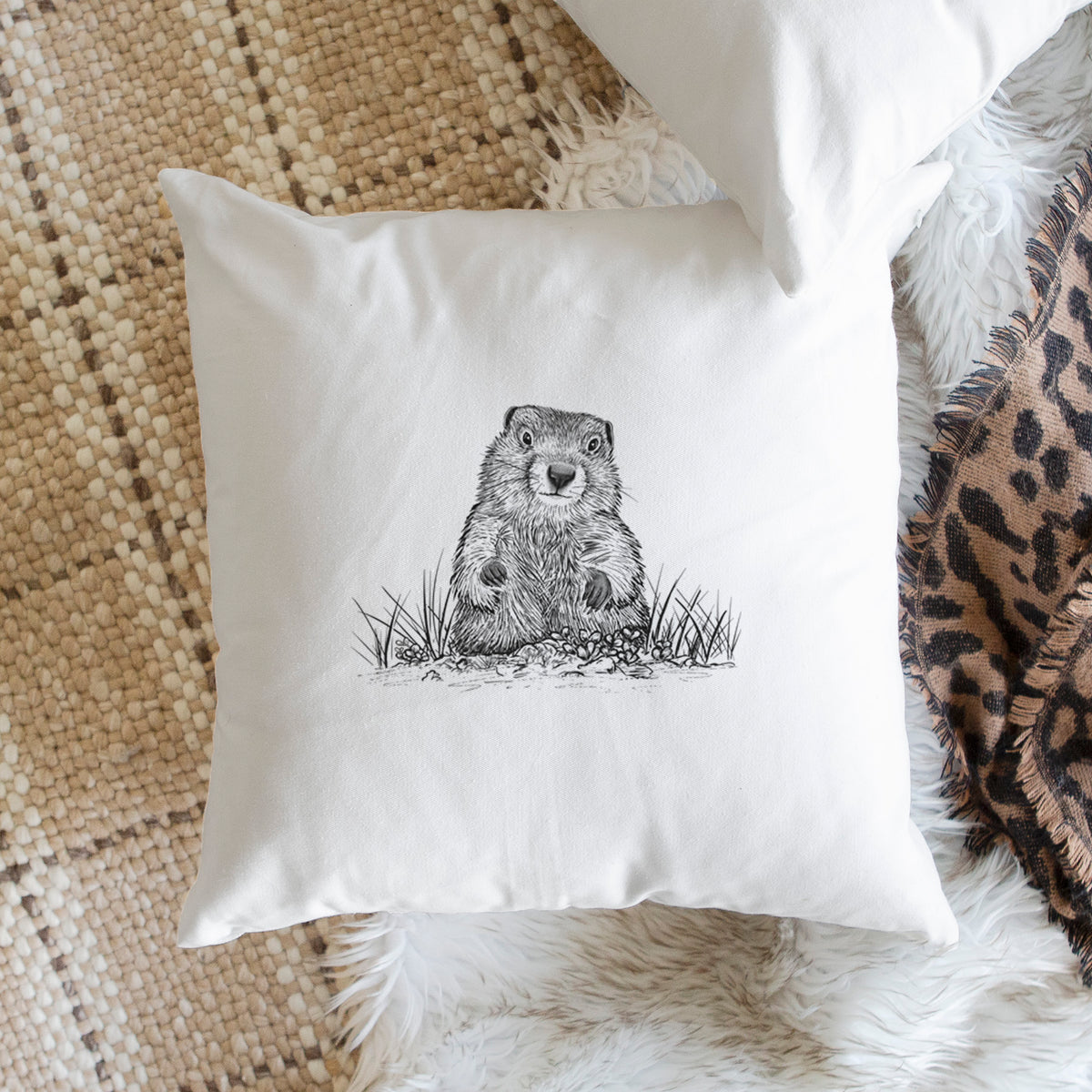 White pillow cover printed with a black ink illustration of a groundhog peeking out of its burrow among grass, on a woven chair.