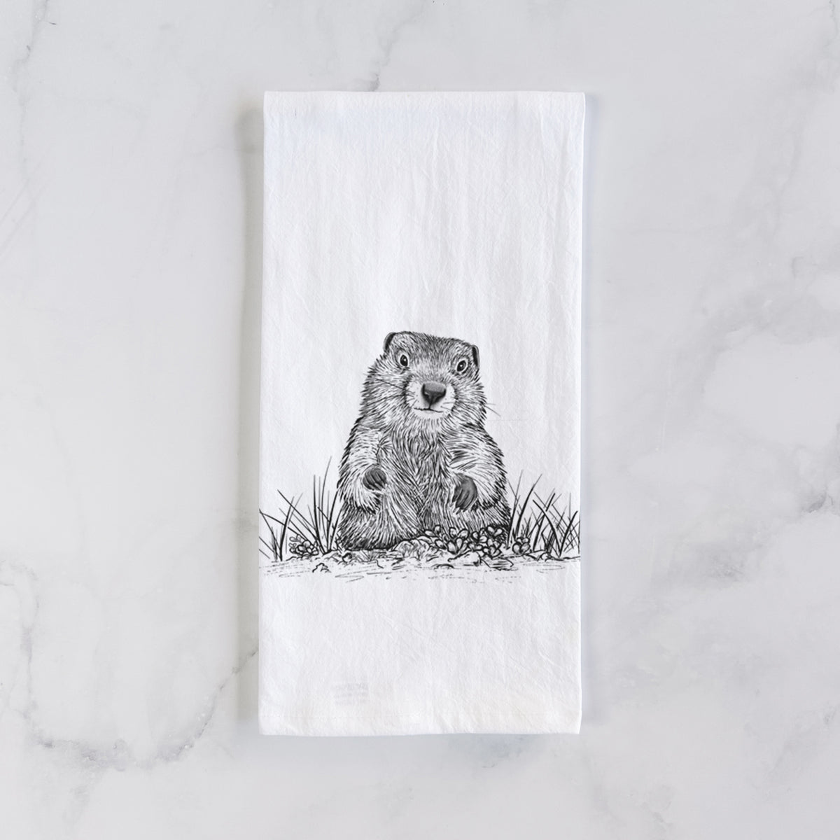 White cotton tea towel laid flat on marble, printed with a black ink illustration of a groundhog peeking out of its burrow among grass.