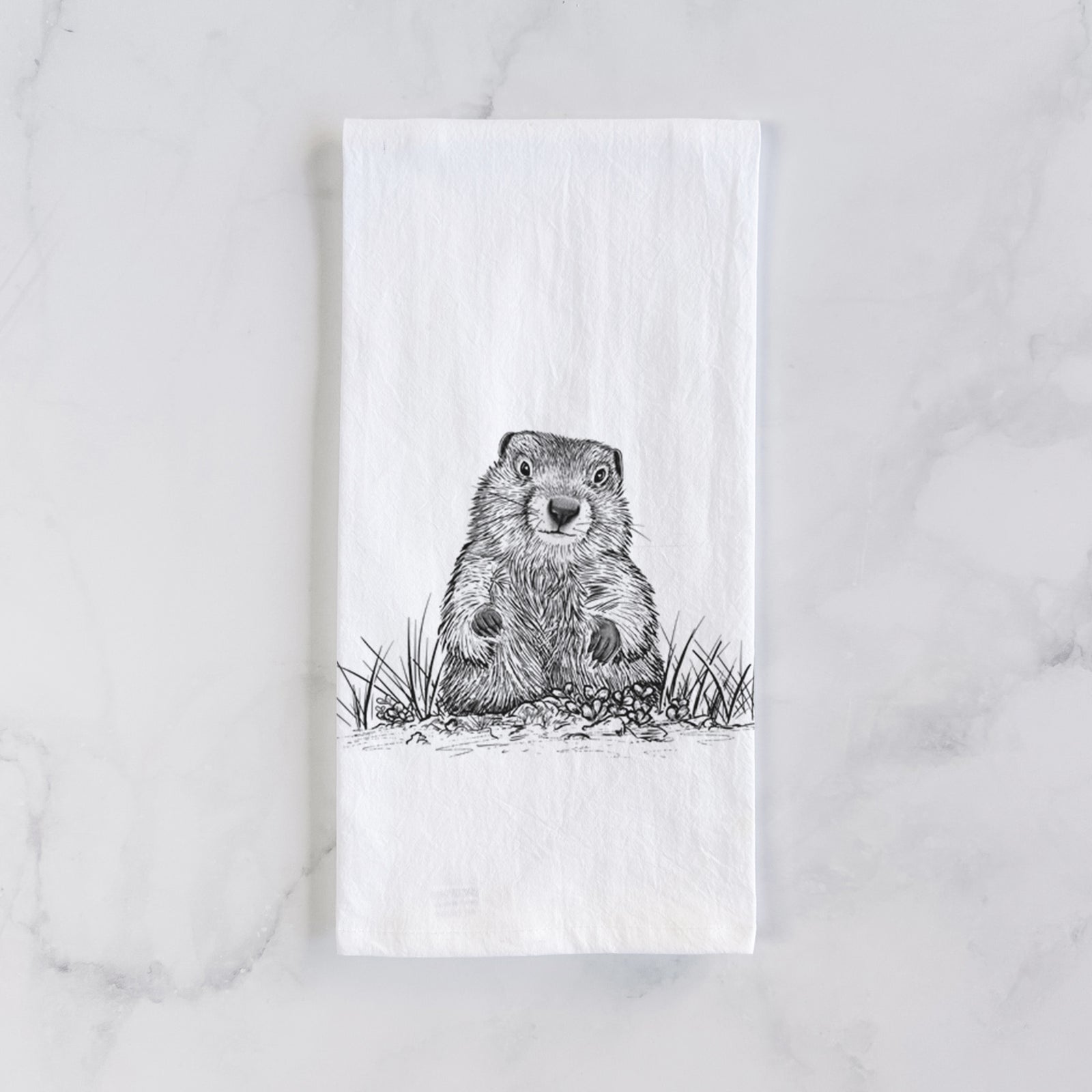 White cotton tea towel printed with a black ink illustration of a groundhog peeking out of its burrow among grass, on a marble counter beside a round wooden cutting board and greenery.