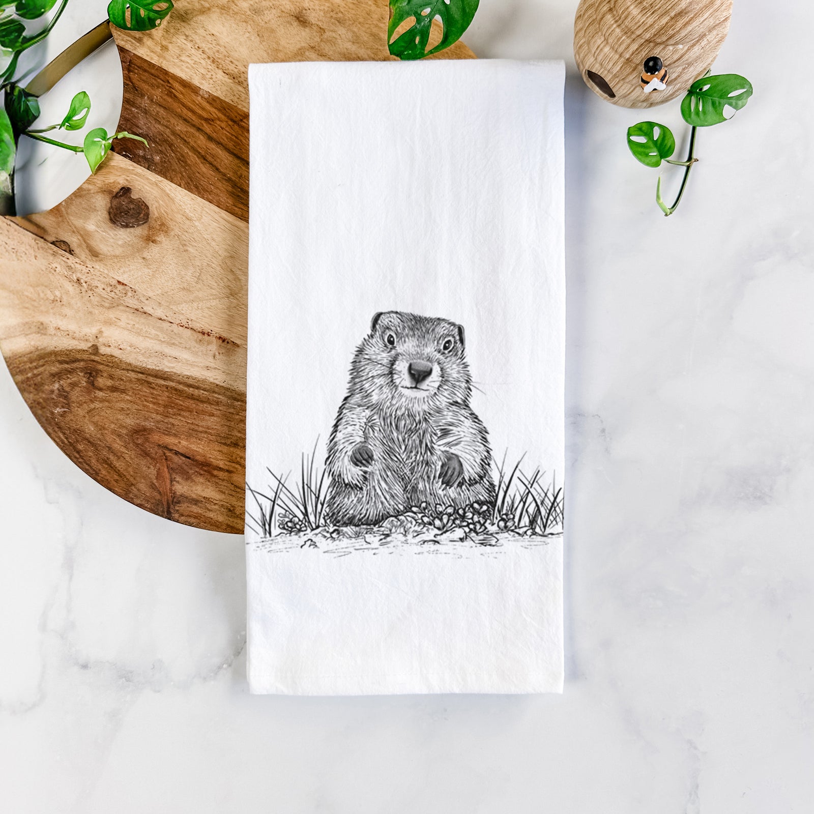 White cotton tea towel printed with a black ink illustration of a groundhog peeking out of its burrow among grass, on a marble counter beside a round wooden cutting board and greenery.