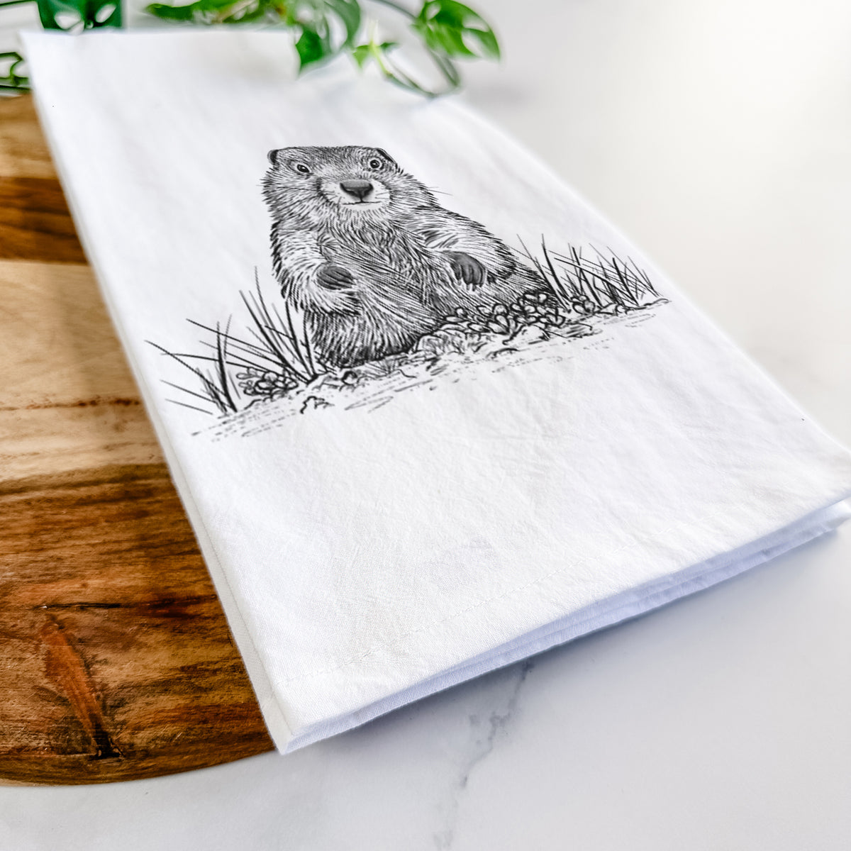 Close-up of a folded white tea towel on a wooden board, showing a black ink illustration of a groundhog peeking out of its burrow among grass.