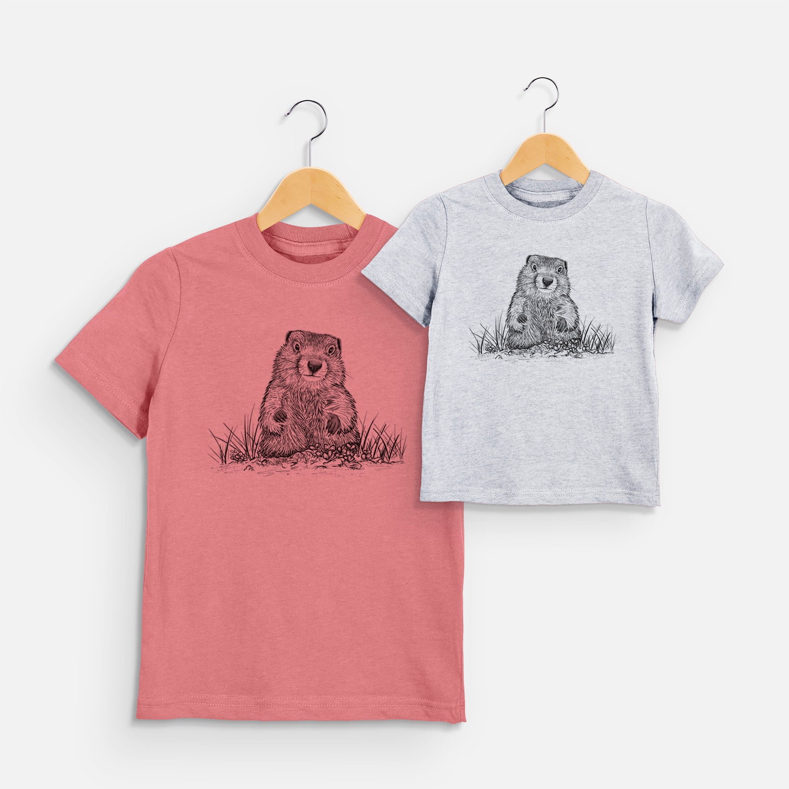 Two youth t-shirts in different colors on hangers, printed with a black ink illustration of a groundhog peeking out of its burrow among grass.