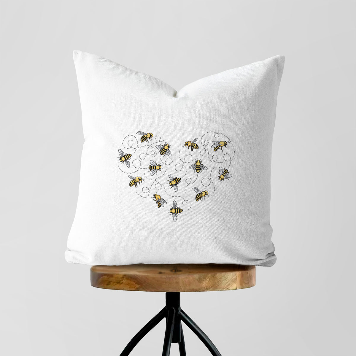 White pillow cover printed with a heart shape made of small bees and looping flight trails, on a wooden stool.