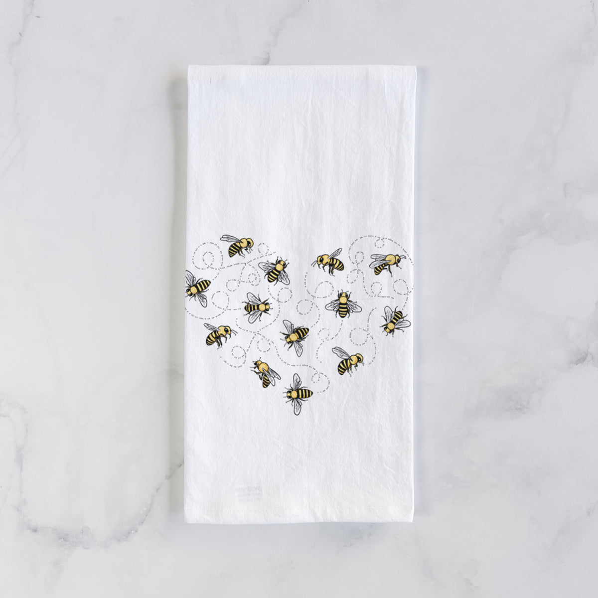 White cotton tea towel laid flat on marble, printed with a heart shape made of small bees and looping flight trails.
