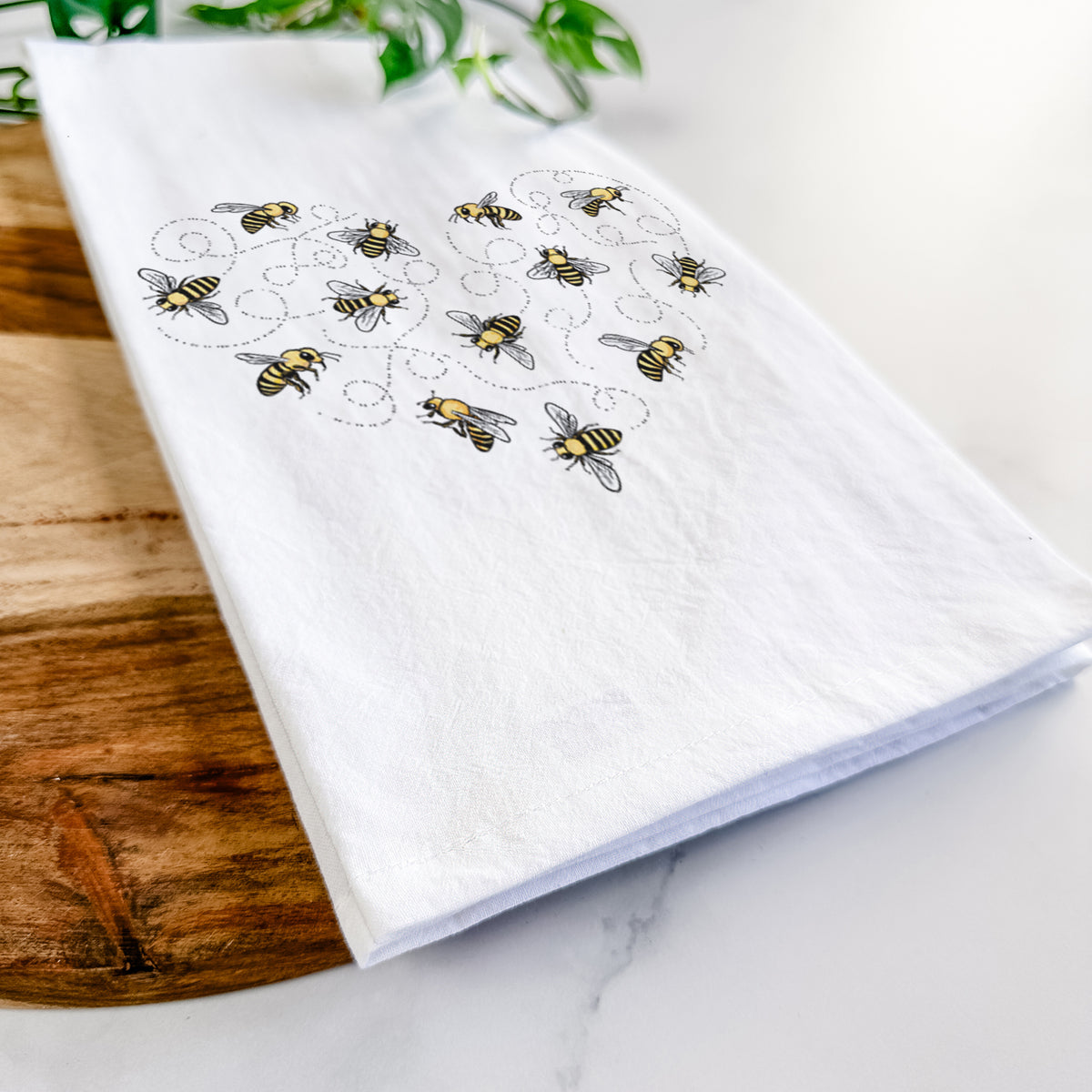 Close-up of a folded white tea towel on a wooden board, showing a heart shape made of small bees and looping flight trails.