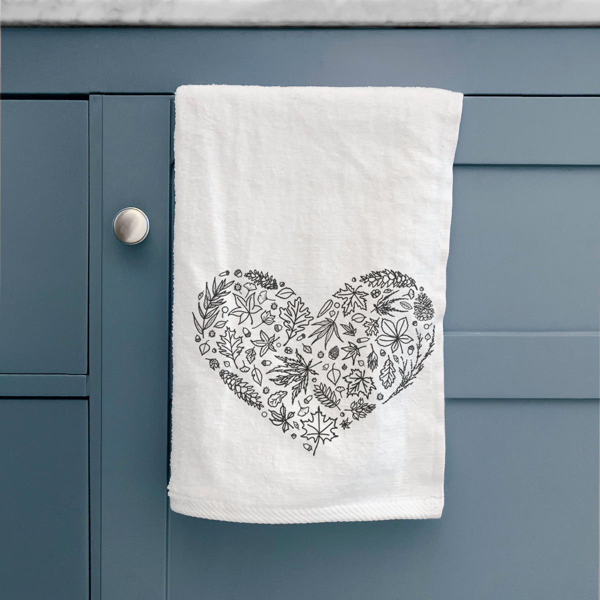 White hand towel printed with a heart shape filled with black ink autumn leaves, hanging on a cabinet door.