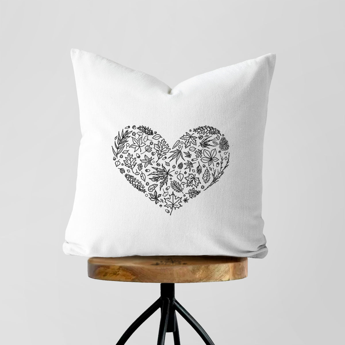 White pillow cover printed with a heart shape filled with black ink autumn leaves, on a wooden stool.