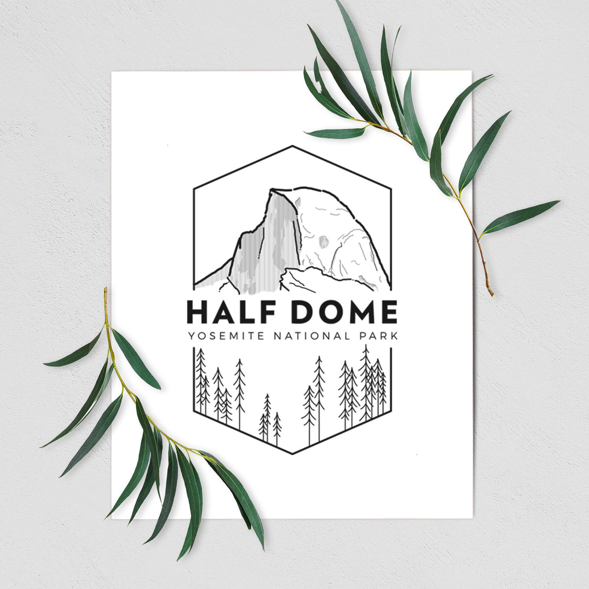 Art print of a black line art badge of Half Dome above a pine forest, with the words Half Dome, Yosemite National Park, shown with eucalyptus leaves.