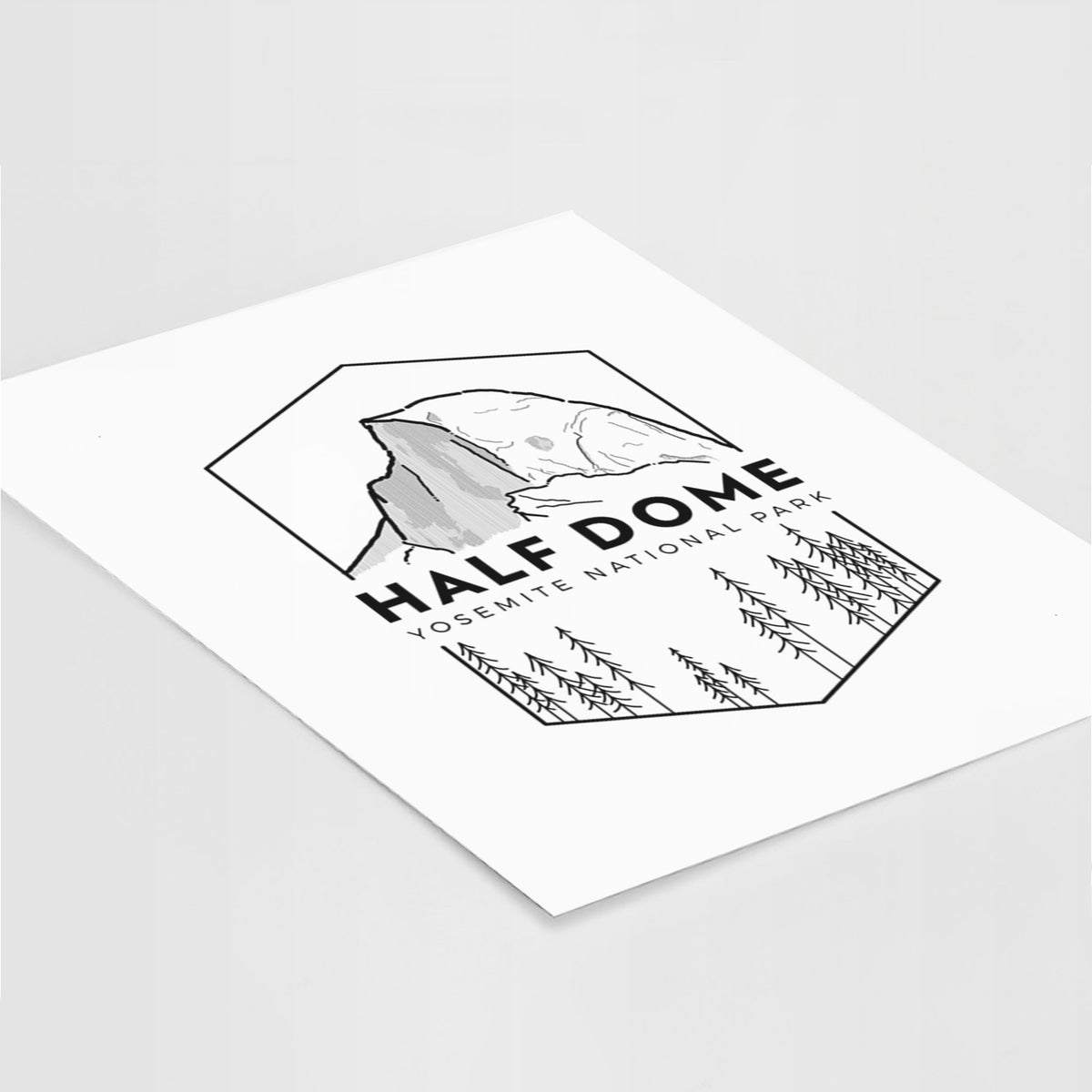 Art print of a black line art badge of Half Dome above a pine forest, with the words Half Dome, Yosemite National Park, lying at an angle on a white surface.