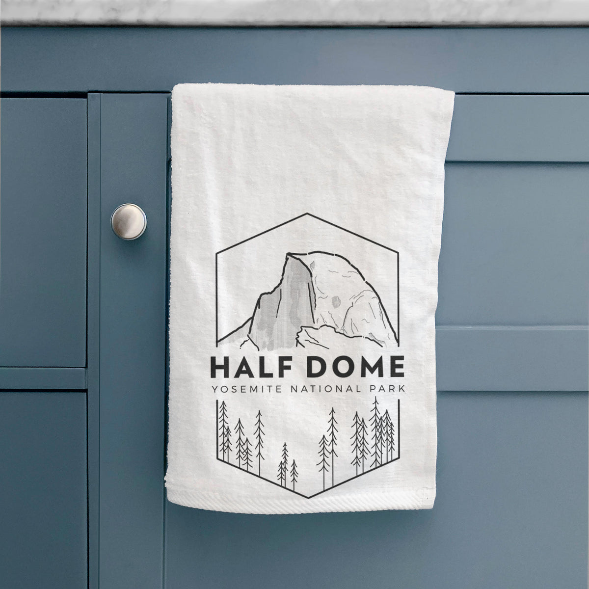 White hand towel printed with a black line art badge of Half Dome above a pine forest, with the words Half Dome, Yosemite National Park, hanging on a cabinet door.