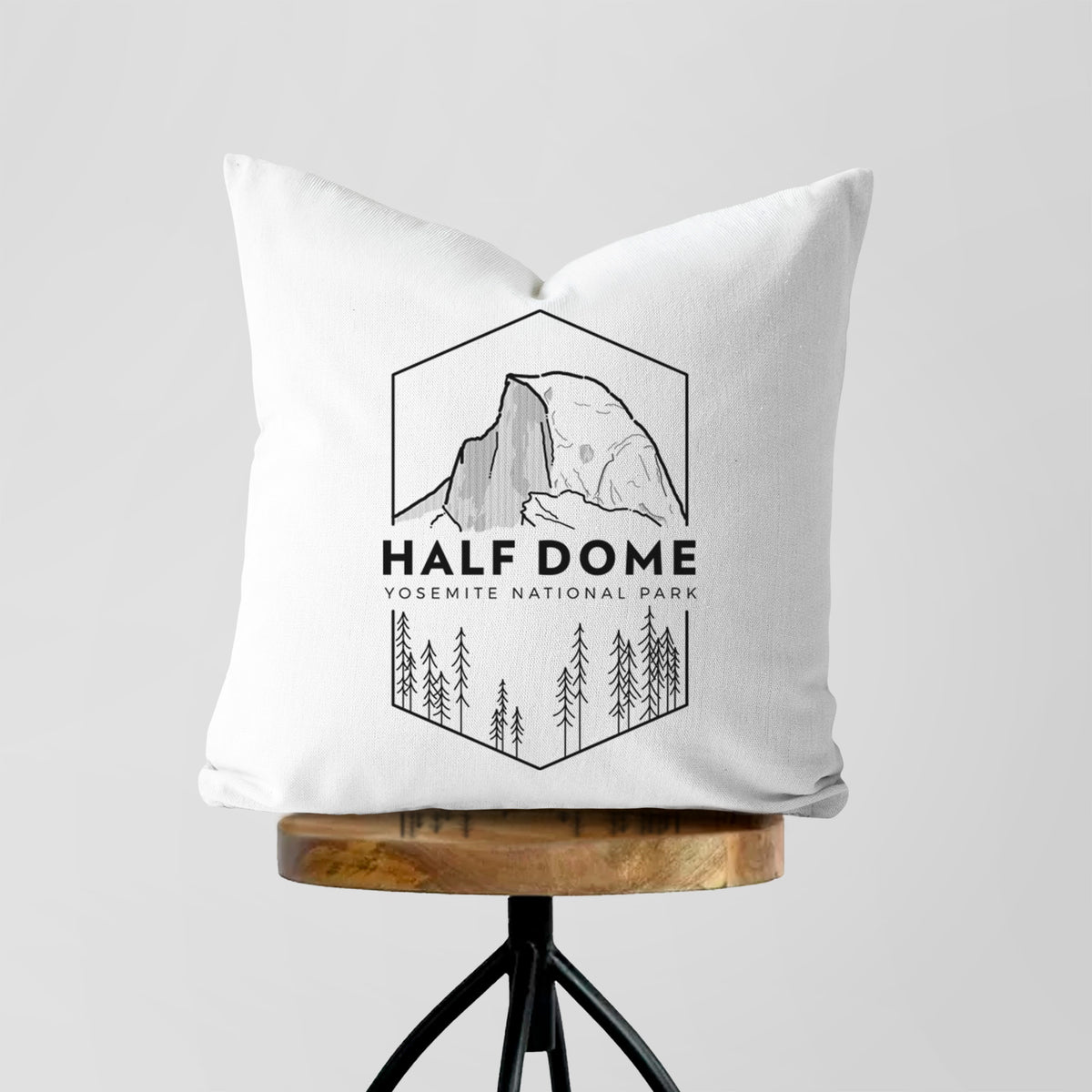 White pillow cover printed with a black line art badge of Half Dome above a pine forest, with the words Half Dome, Yosemite National Park, on a wooden stool.