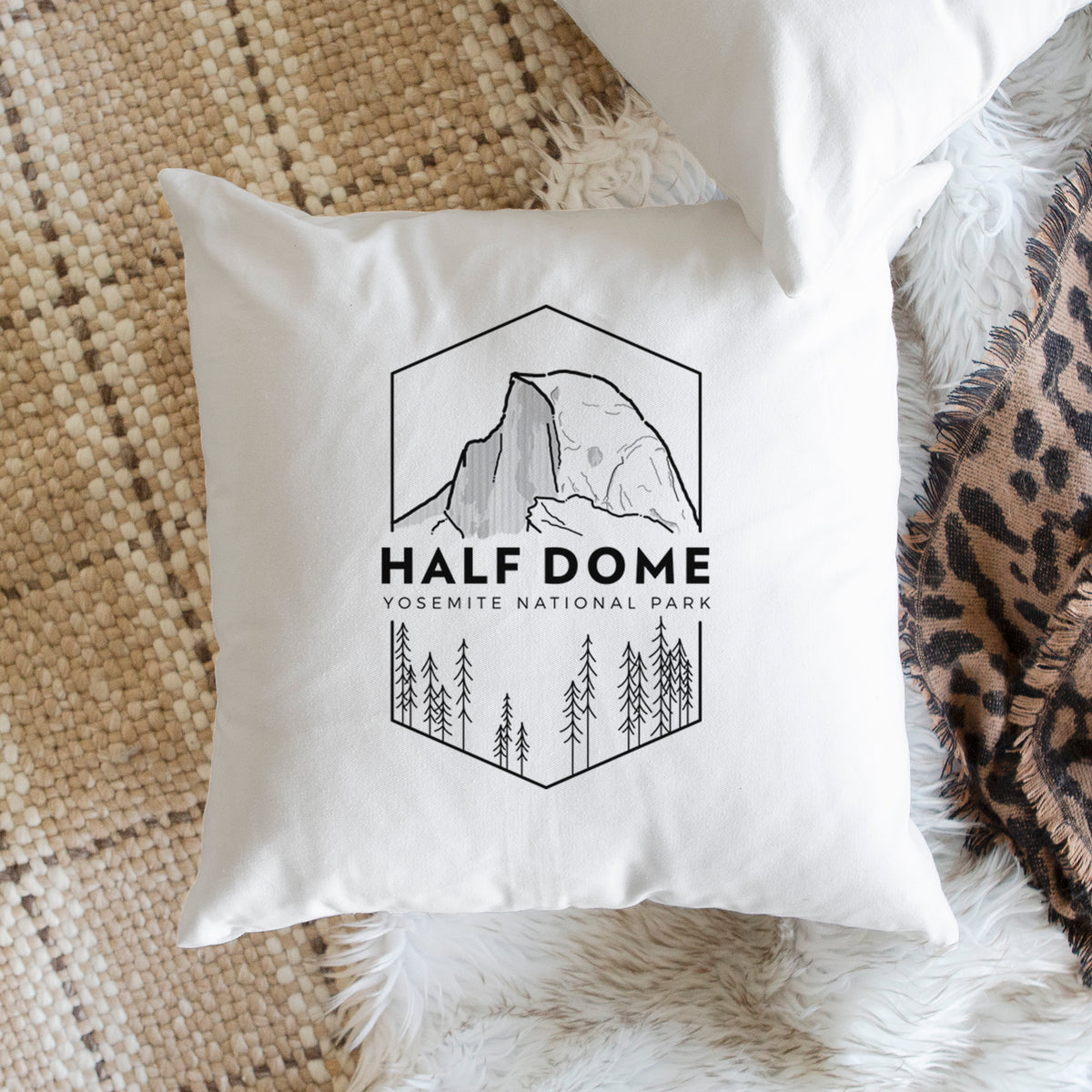 White pillow cover printed with a black line art badge of Half Dome above a pine forest, with the words Half Dome, Yosemite National Park, on a woven chair.