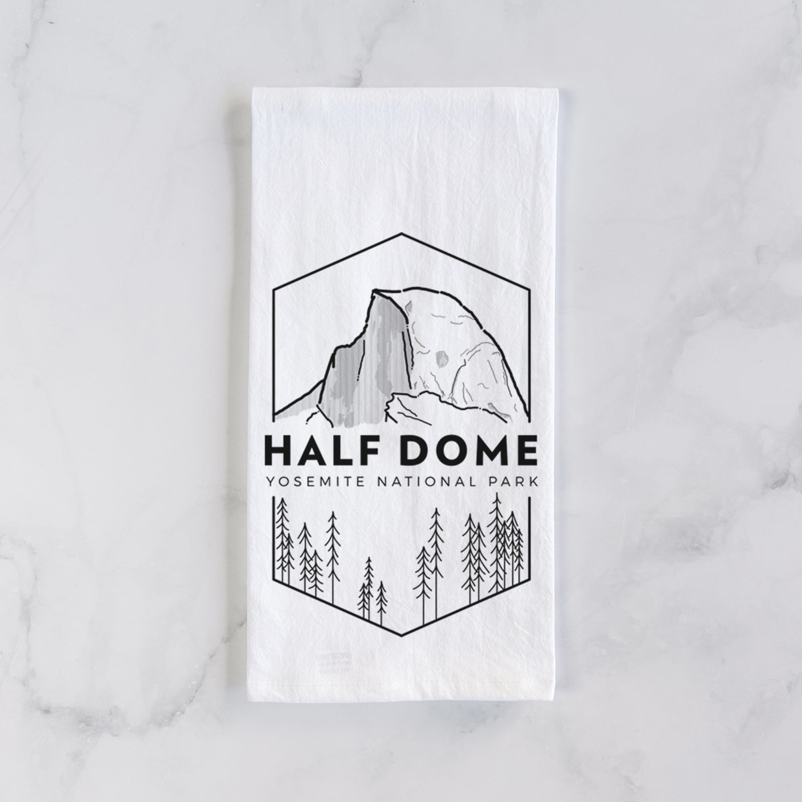 White cotton tea towel printed with a black line art badge of Half Dome above a pine forest, with the words Half Dome, Yosemite National Park, on a marble counter beside a round wooden cutting board and greenery.