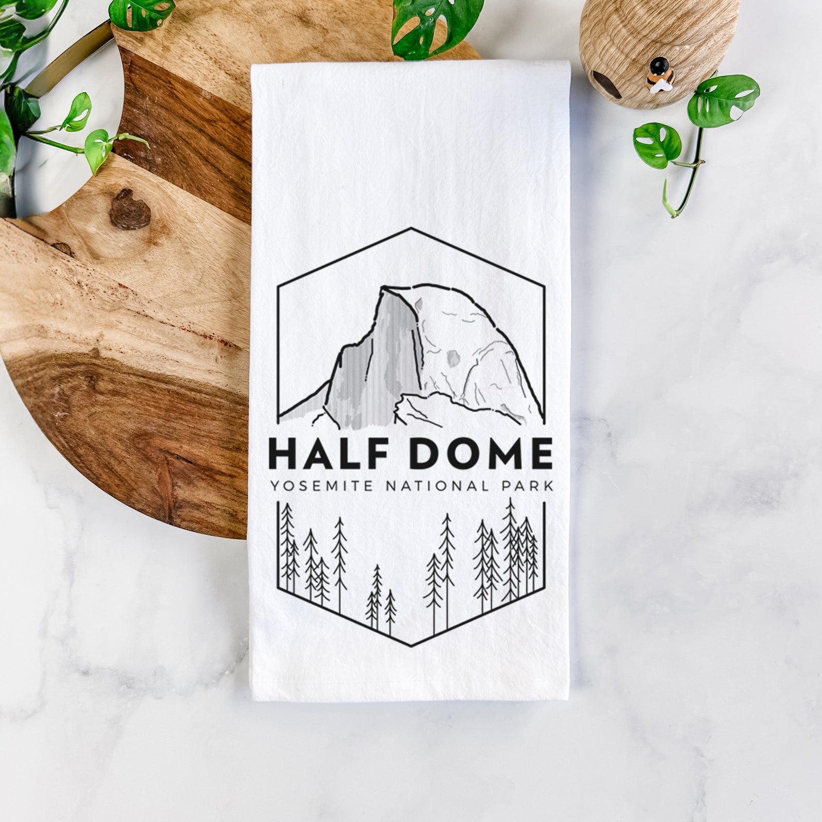 White cotton tea towel printed with a black line art badge of Half Dome above a pine forest, with the words Half Dome, Yosemite National Park, on a marble counter beside a round wooden cutting board and greenery.