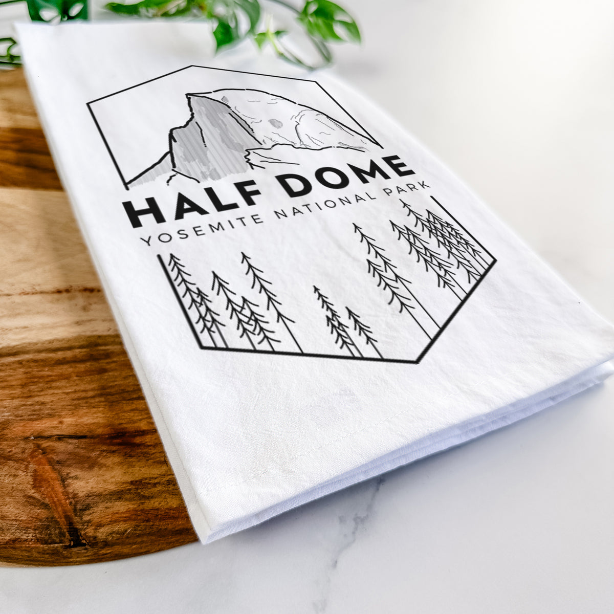 Close-up of a folded white tea towel on a wooden board, showing a black line art badge of Half Dome above a pine forest, with the words Half Dome, Yosemite National Park.