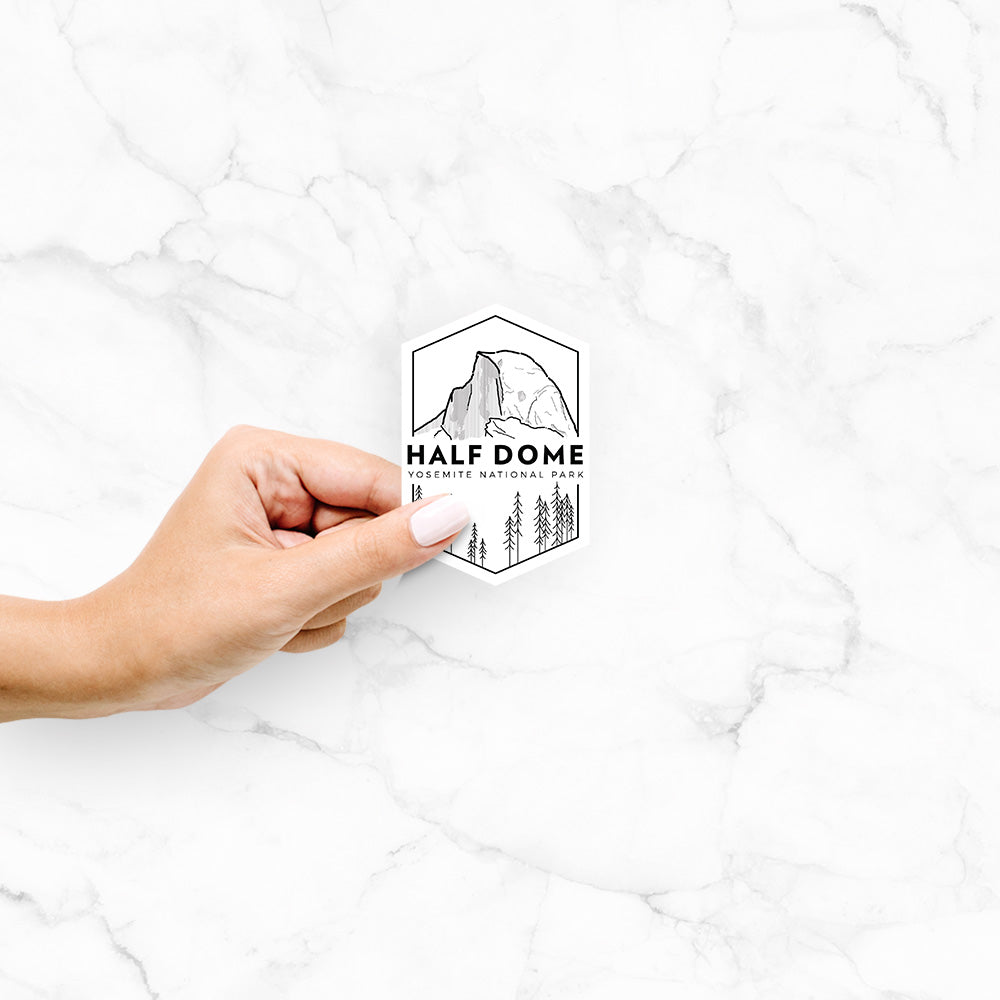 Hand holding a 4 inch vinyl decal of a black line art badge of Half Dome above a pine forest, with the words Half Dome, Yosemite National Park.