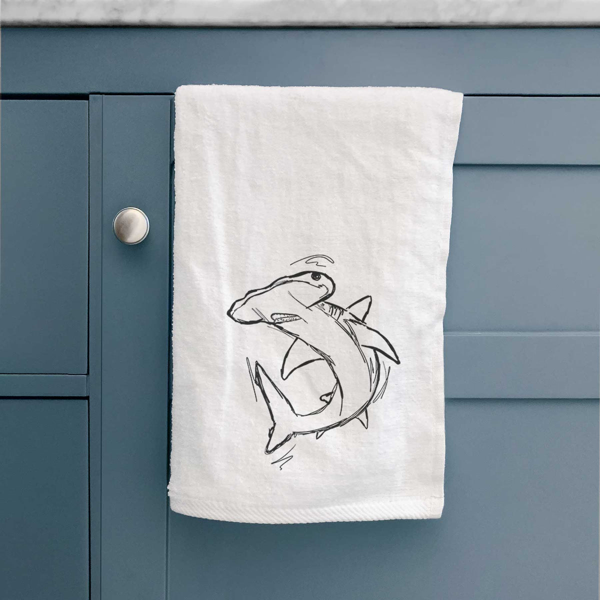 White hand towel printed with a black ink illustration of a hammerhead shark swimming in a curve, hanging on a cabinet door.