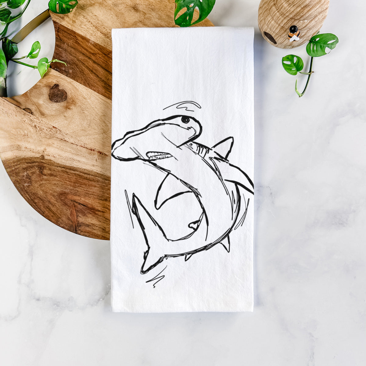 White cotton tea towel printed with a black ink illustration of a hammerhead shark swimming in a curve, on a marble counter beside a round wooden cutting board and greenery.