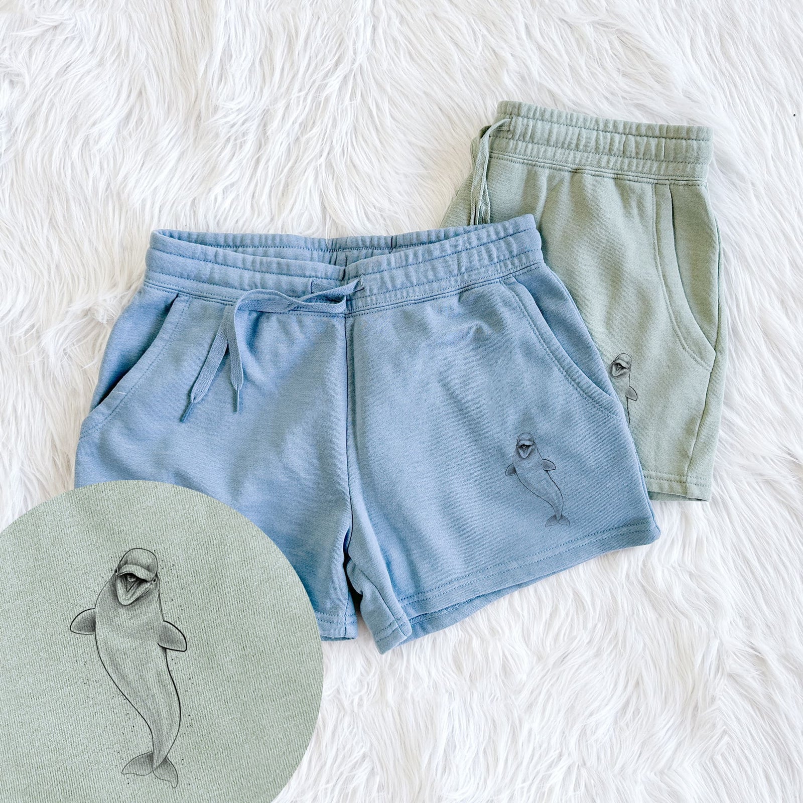 Two pairs of Cali Wave shorts in different colors printed with a black ink illustration of a smiling beluga whale swimming upward, with a close-up of the print.