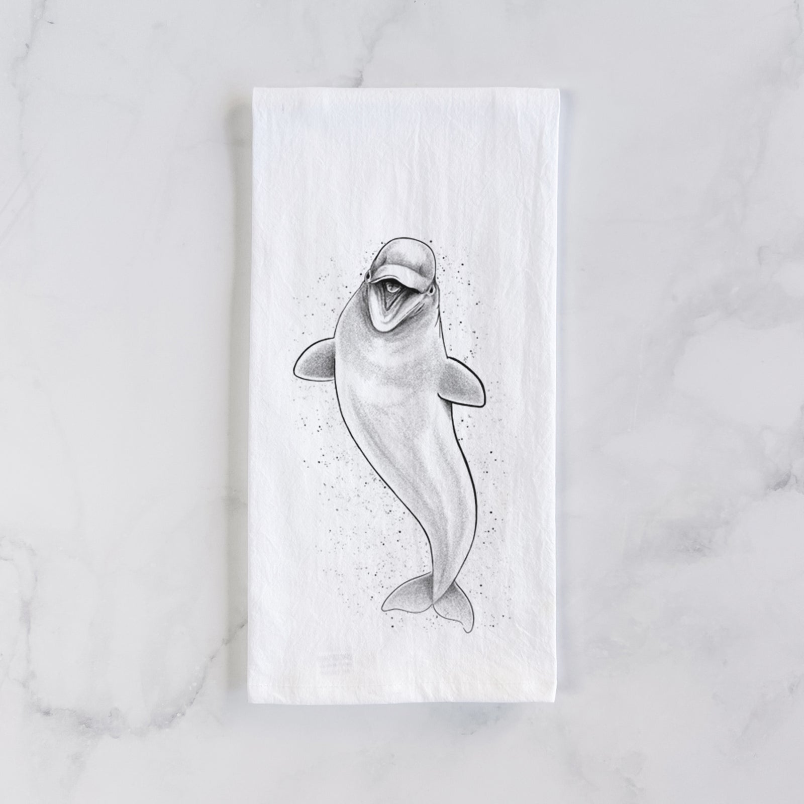 White cotton tea towel printed with a black ink illustration of a smiling beluga whale swimming upward, on a marble counter beside a round wooden cutting board and greenery.