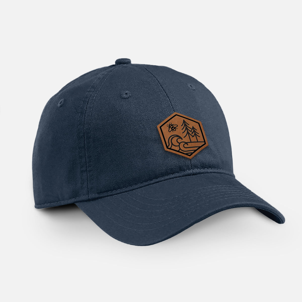 Navy cotton baseball hat with a brown leather hexagon patch engraved with a bee, ocean waves and pine trees on the front.