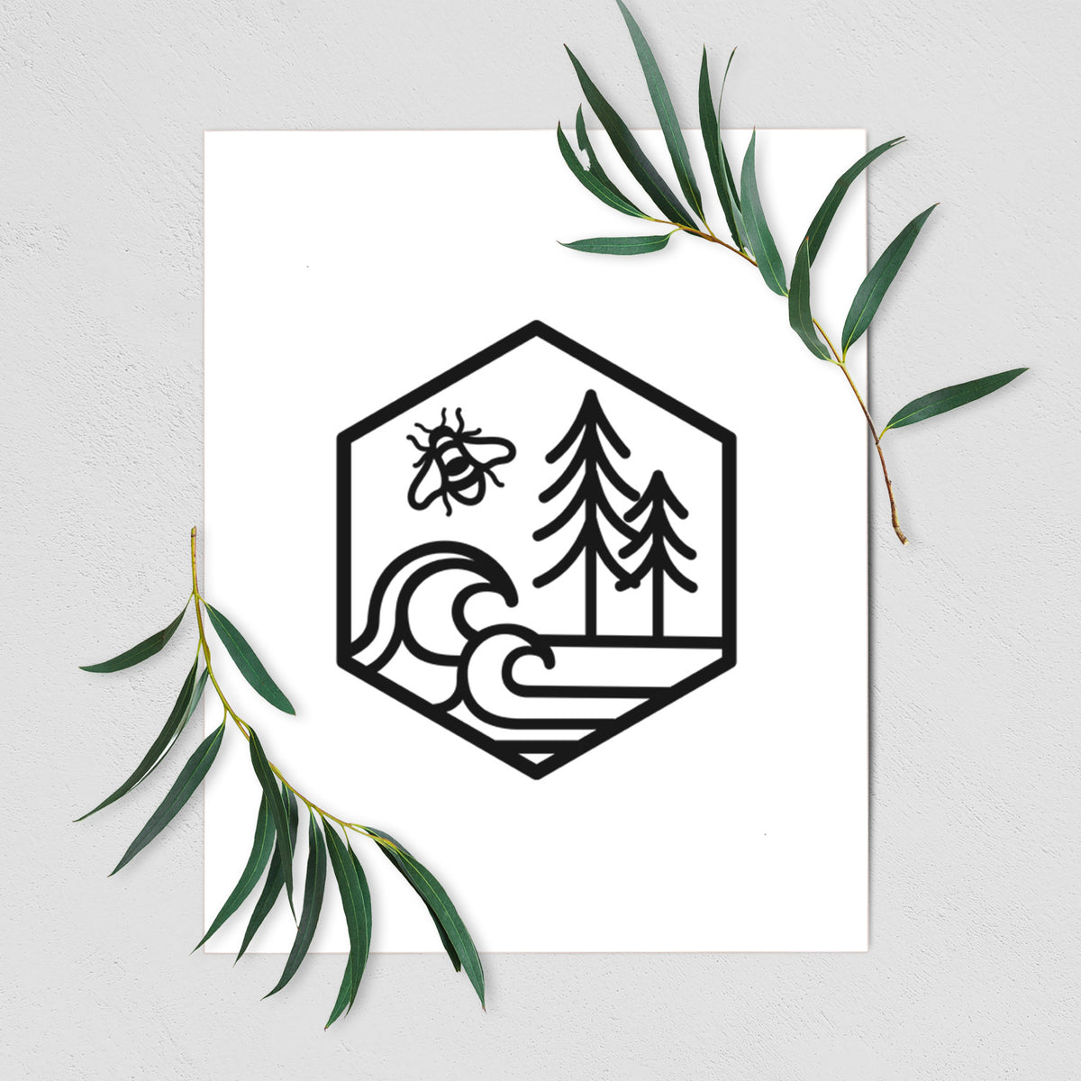 Art print of a black line art hexagon holding a bee, ocean waves and pine trees, shown with eucalyptus leaves.