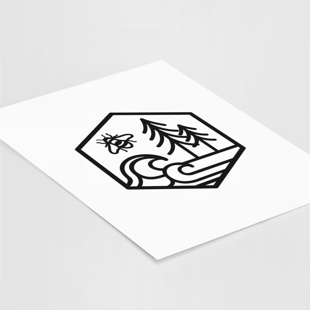 Art print of a black line art hexagon holding a bee, ocean waves and pine trees, lying at an angle on a white surface.