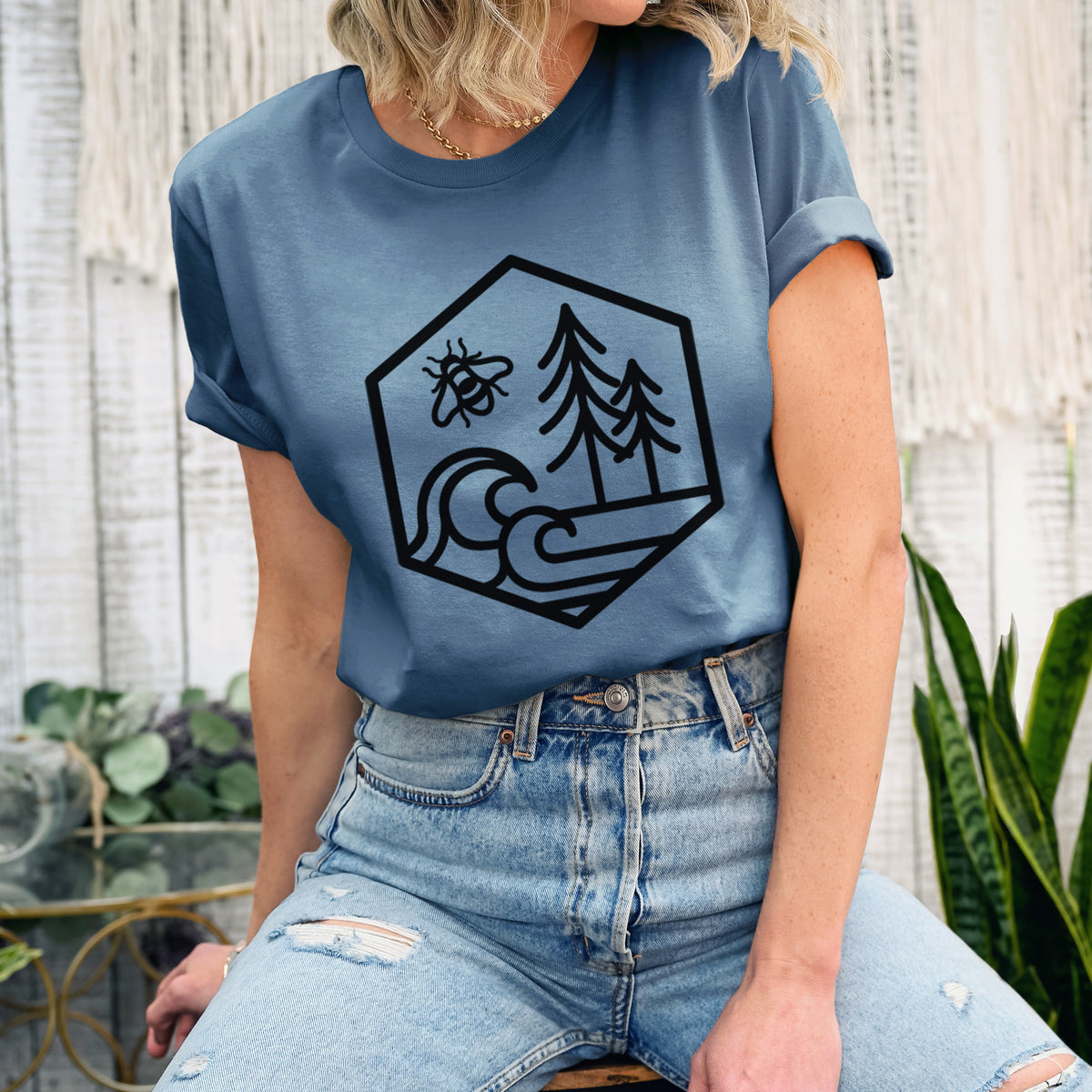 Woman wearing a steel blue lightweight cotton t-shirt printed with a black line art hexagon holding a bee, ocean waves and pine trees.