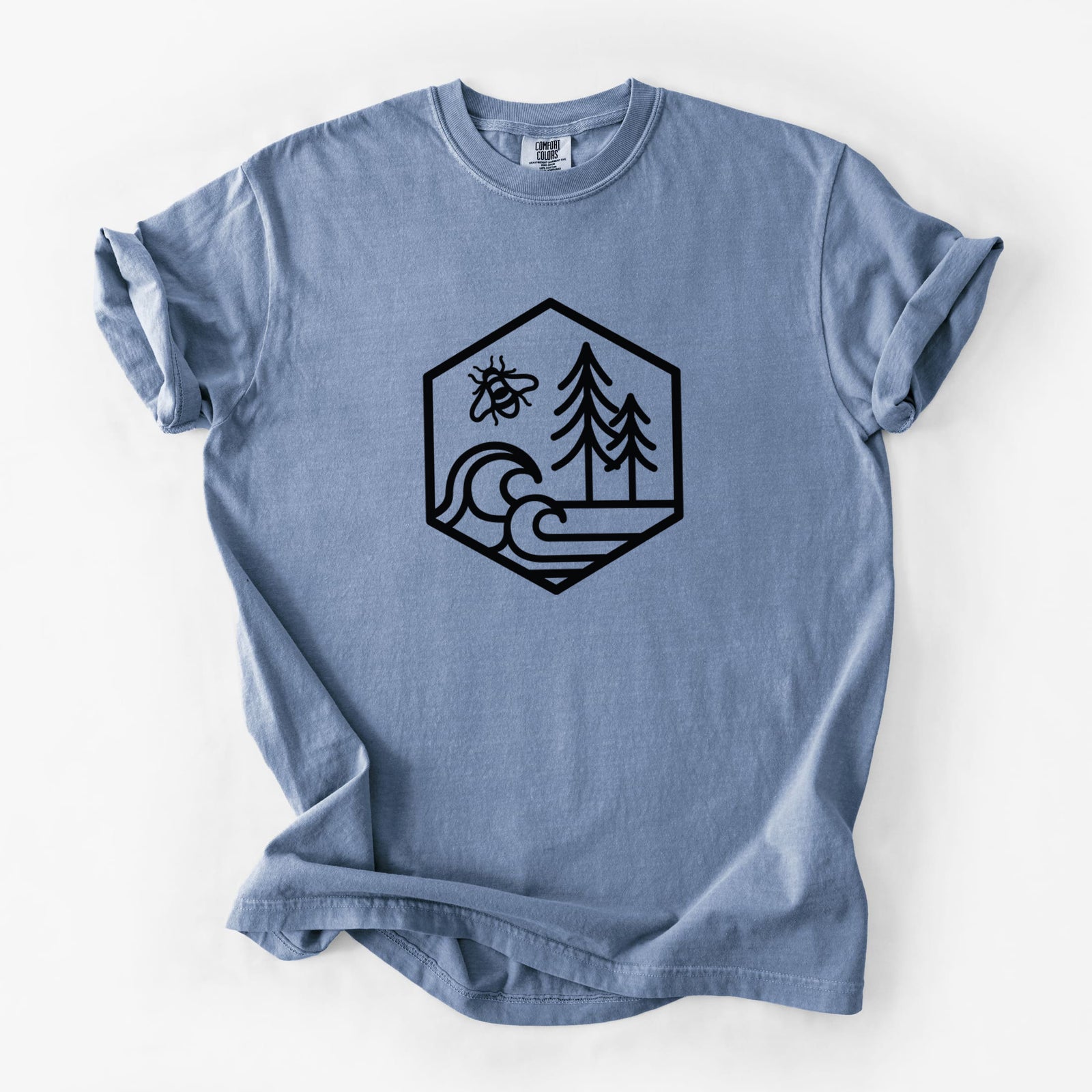 Blue jean Comfort Colors t-shirt laid flat, printed with a black line art hexagon holding a bee, ocean waves and pine trees.