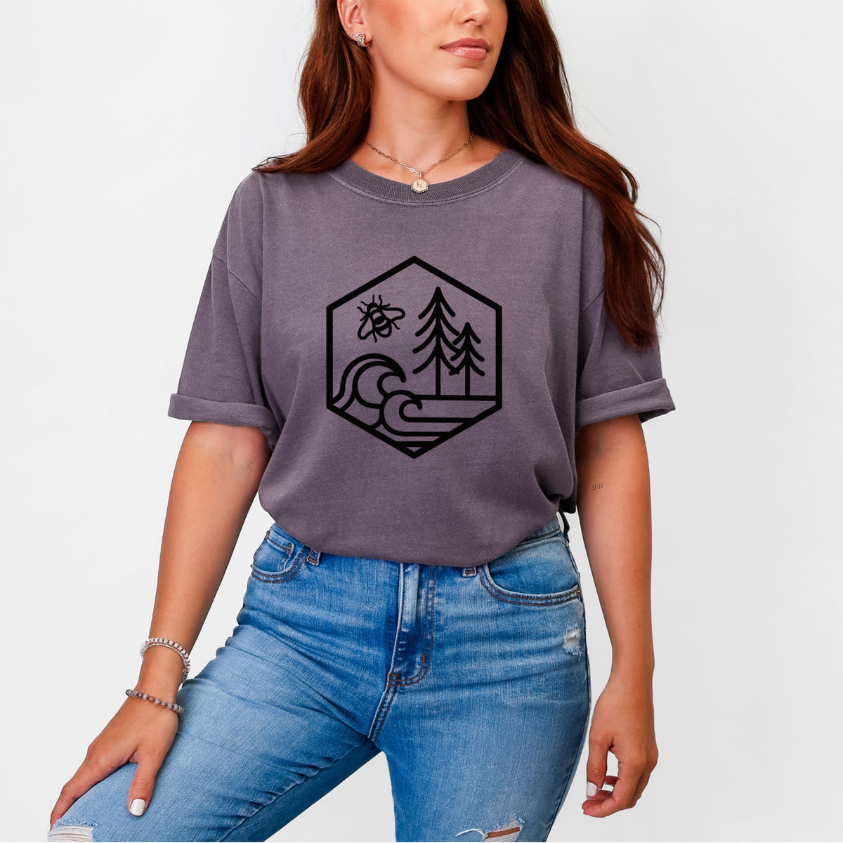 Woman wearing a Comfort Colors t-shirt printed with a black line art hexagon holding a bee, ocean waves and pine trees.