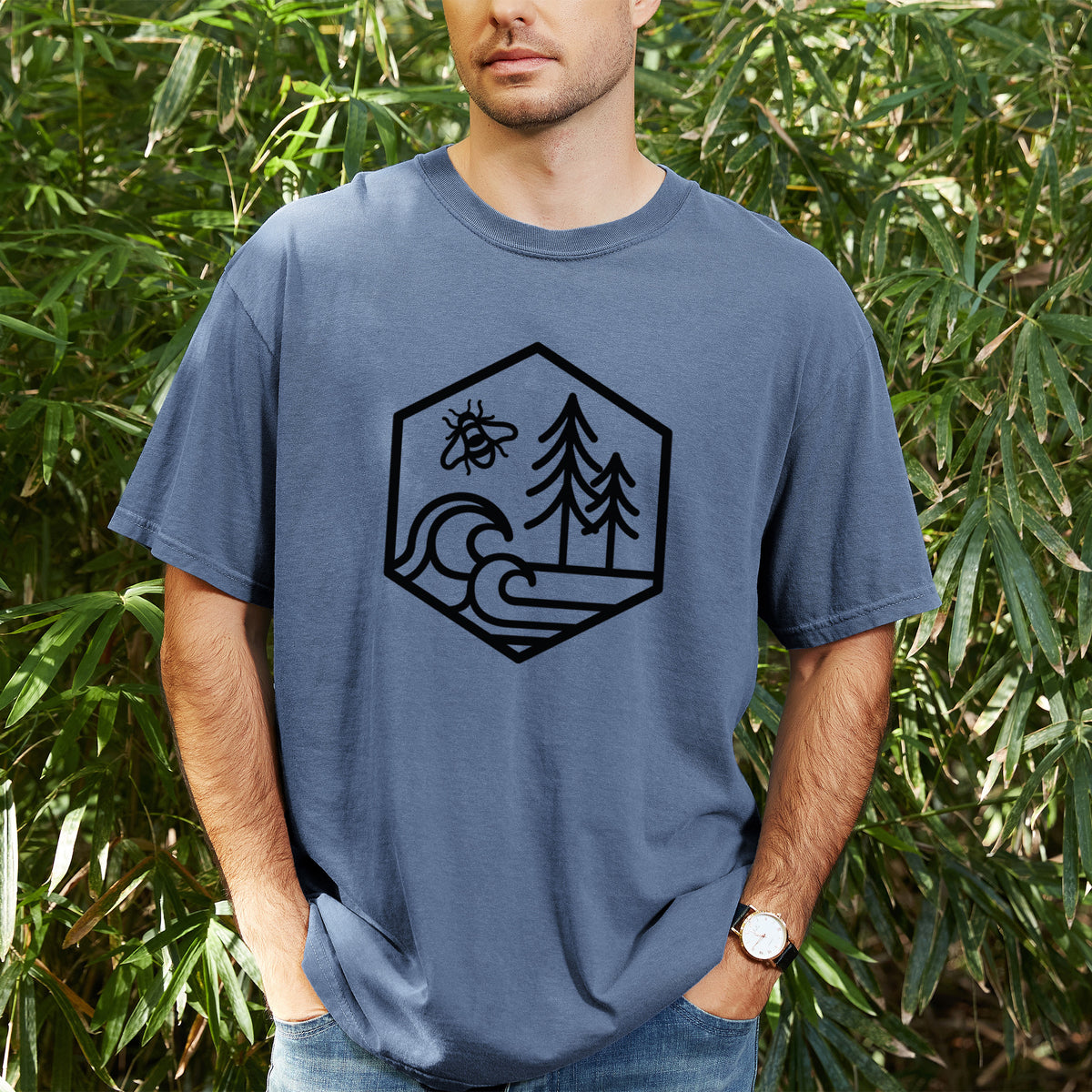 Man wearing a Comfort Colors t-shirt printed with a black line art hexagon holding a bee, ocean waves and pine trees, outdoors.