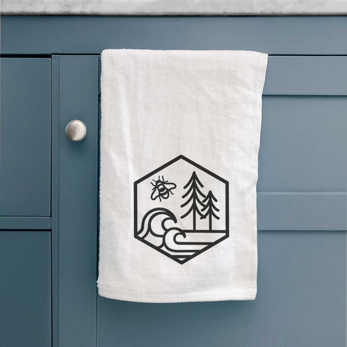 White hand towel printed with a black line art hexagon holding a bee, ocean waves and pine trees, hanging on a cabinet door.