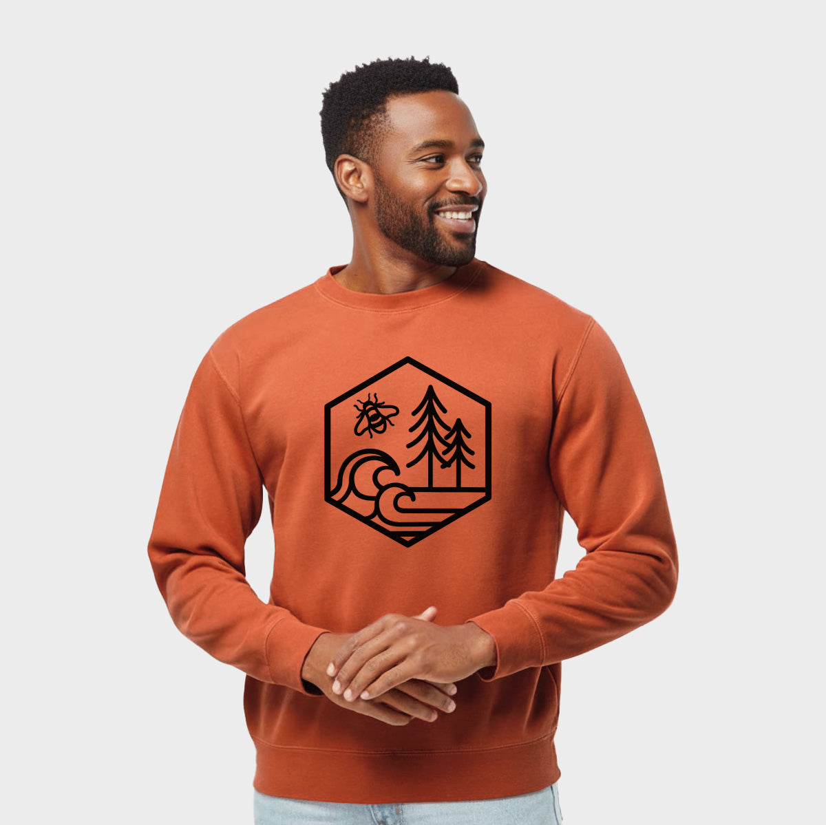 Model wearing an amber orange pigment dyed crewneck sweatshirt printed with a black line art hexagon holding a bee, ocean waves and pine trees.