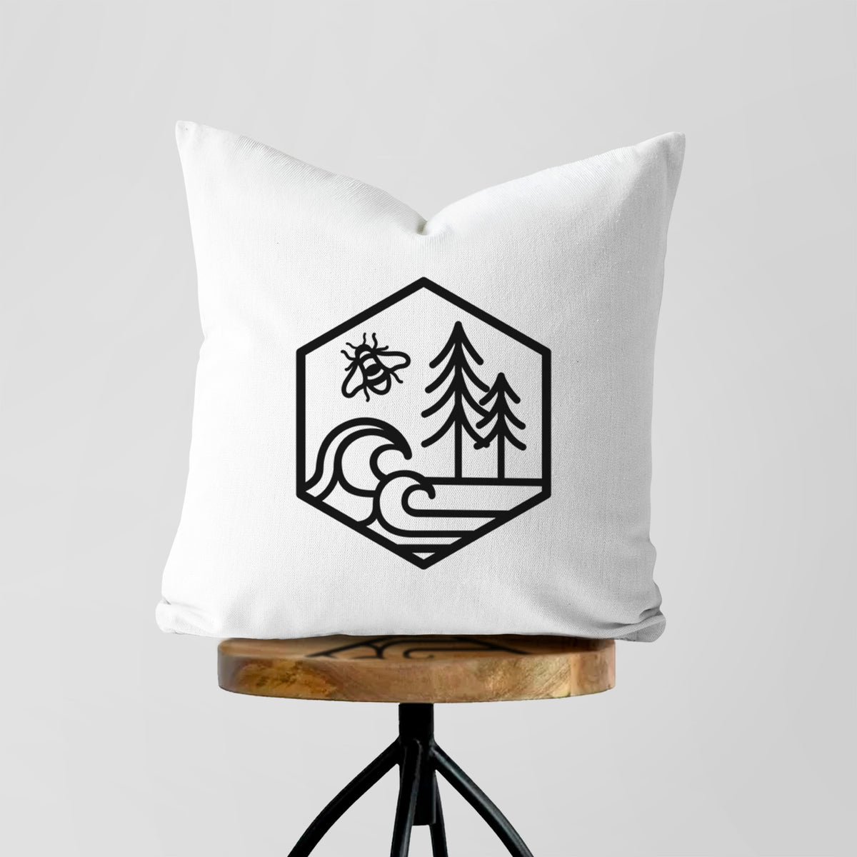 White pillow cover printed with a black line art hexagon holding a bee, ocean waves and pine trees, on a wooden stool.