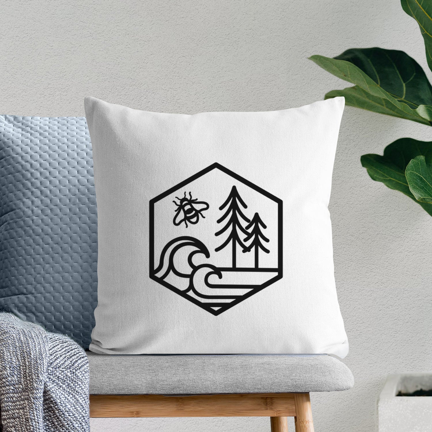 White pillow cover printed with a black line art hexagon holding a bee, ocean waves and pine trees, on a bench beside plants.