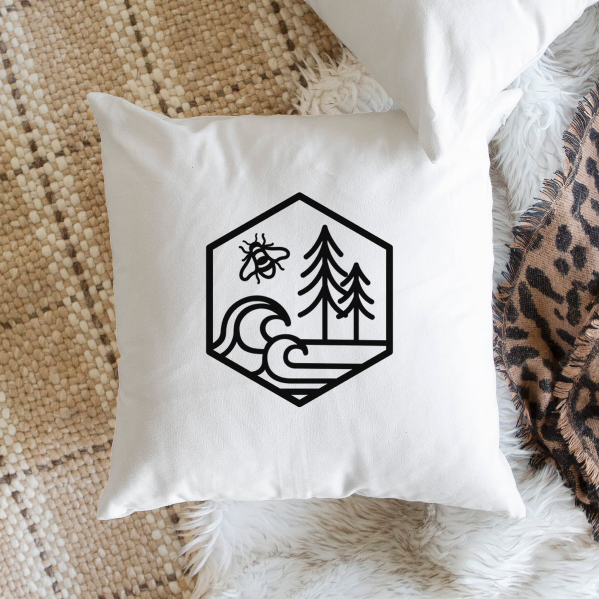 White pillow cover printed with a black line art hexagon holding a bee, ocean waves and pine trees, on a woven chair.