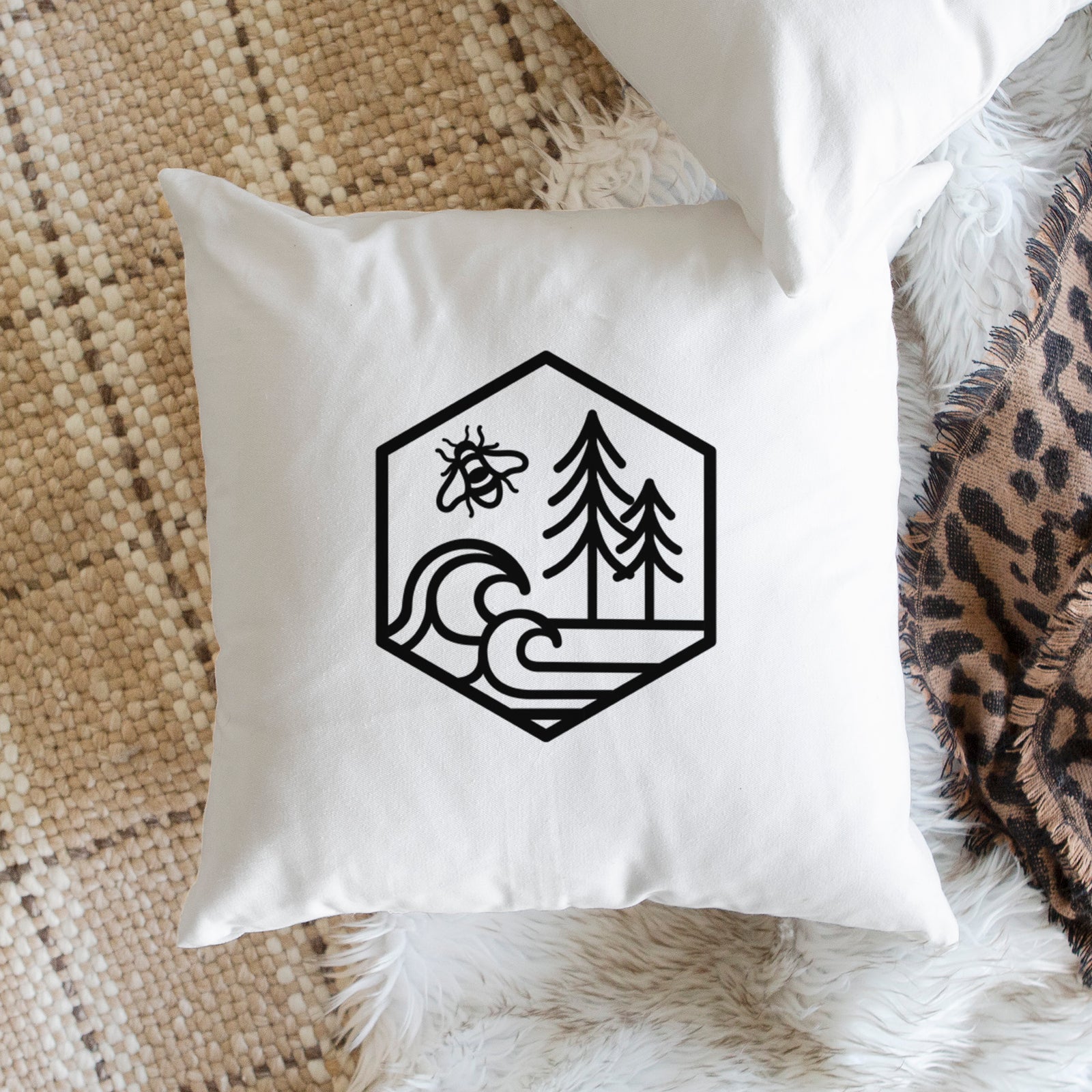 White pillow cover printed with a black line art hexagon holding a bee, ocean waves and pine trees, on a bench beside plants.