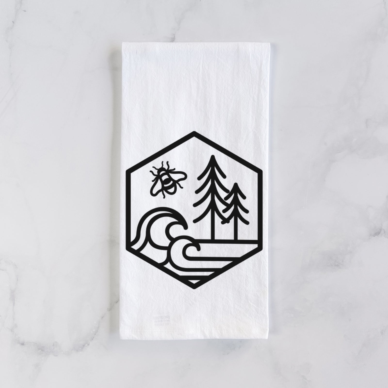 White cotton tea towel printed with a black line art hexagon holding a bee, ocean waves and pine trees, on a marble counter beside a round wooden cutting board and greenery.