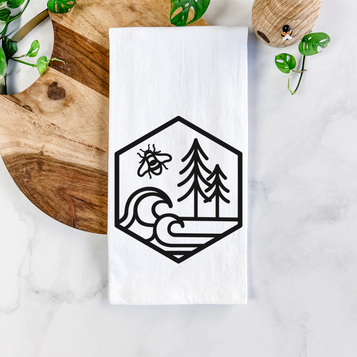 White cotton tea towel printed with a black line art hexagon holding a bee, ocean waves and pine trees, on a marble counter beside a round wooden cutting board and greenery.