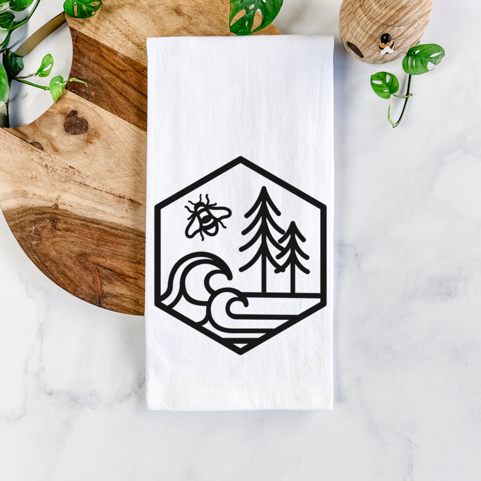 White cotton tea towel printed with a black line art hexagon holding a bee, ocean waves and pine trees, on a marble counter beside a round wooden cutting board and greenery.
