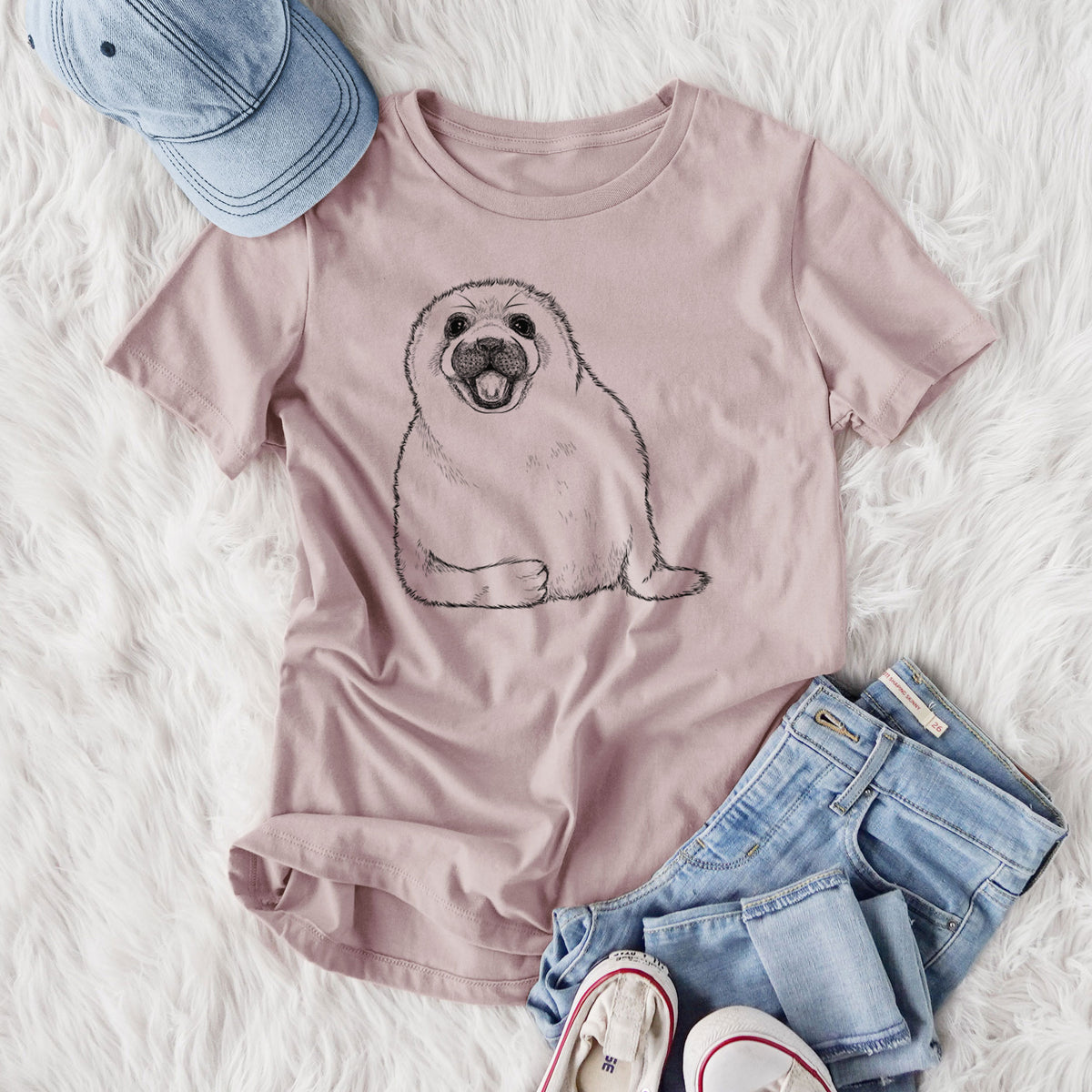 Women's relaxed fit cotton t-shirt printed with a black ink illustration of a smiling harp seal pup lying down, styled flat on a white faux fur rug with jeans.