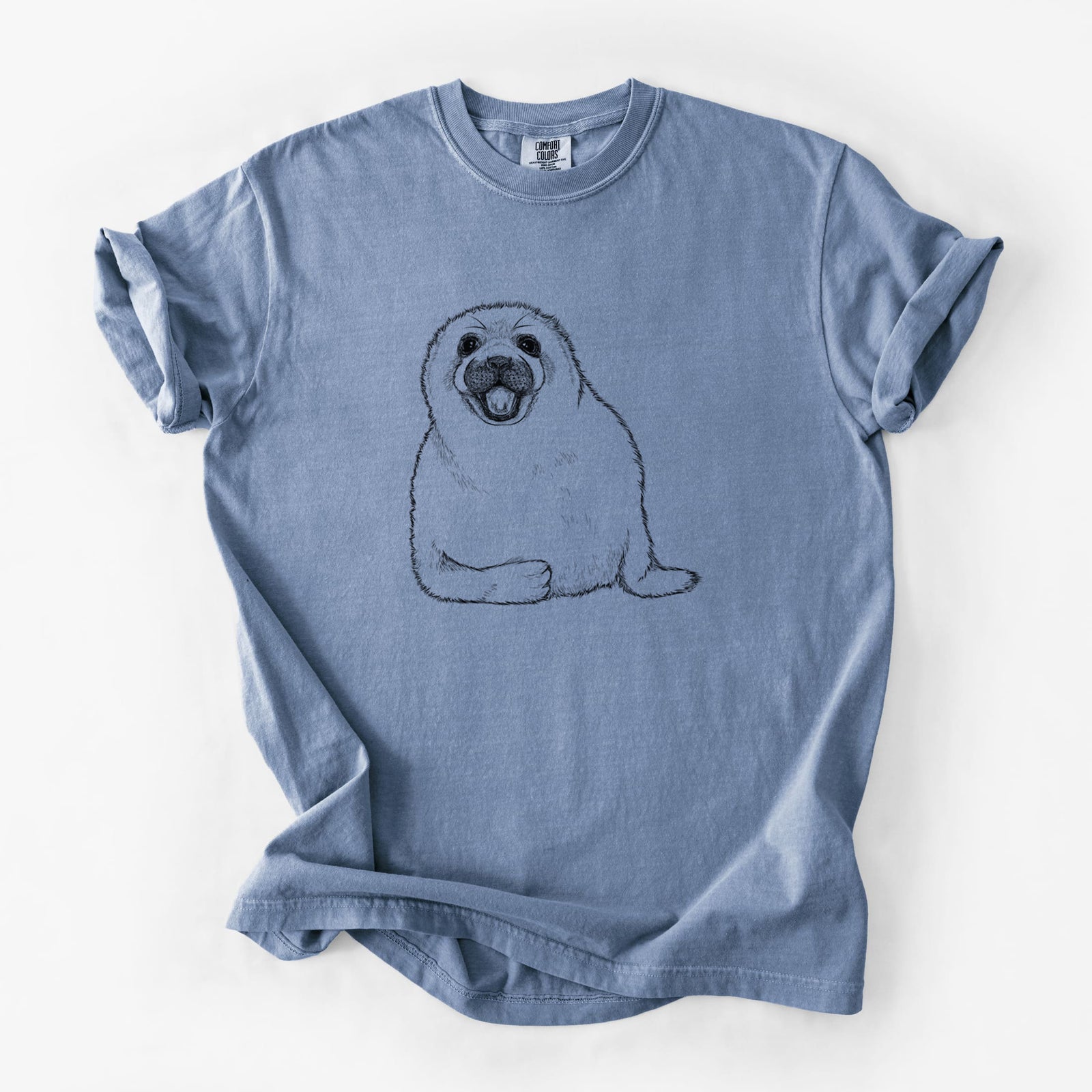Blue jean Comfort Colors t-shirt laid flat, printed with a black ink illustration of a smiling harp seal pup lying down.