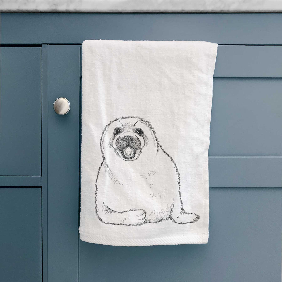 White hand towel printed with a black ink illustration of a smiling harp seal pup lying down, hanging on a cabinet door.
