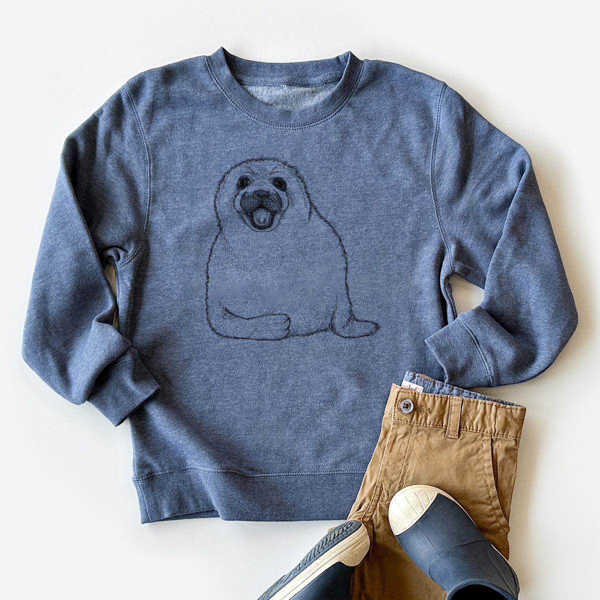 Youth crewneck sweatshirt printed with a black ink illustration of a smiling harp seal pup lying down, styled flat with khaki pants and shoes.