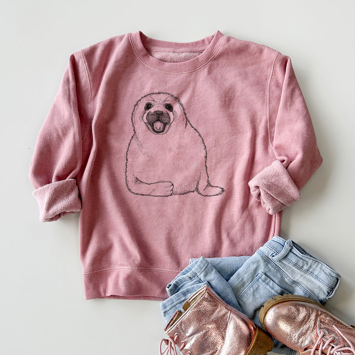 Youth crewneck sweatshirt printed with a black ink illustration of a smiling harp seal pup lying down, styled flat with jeans and boots.