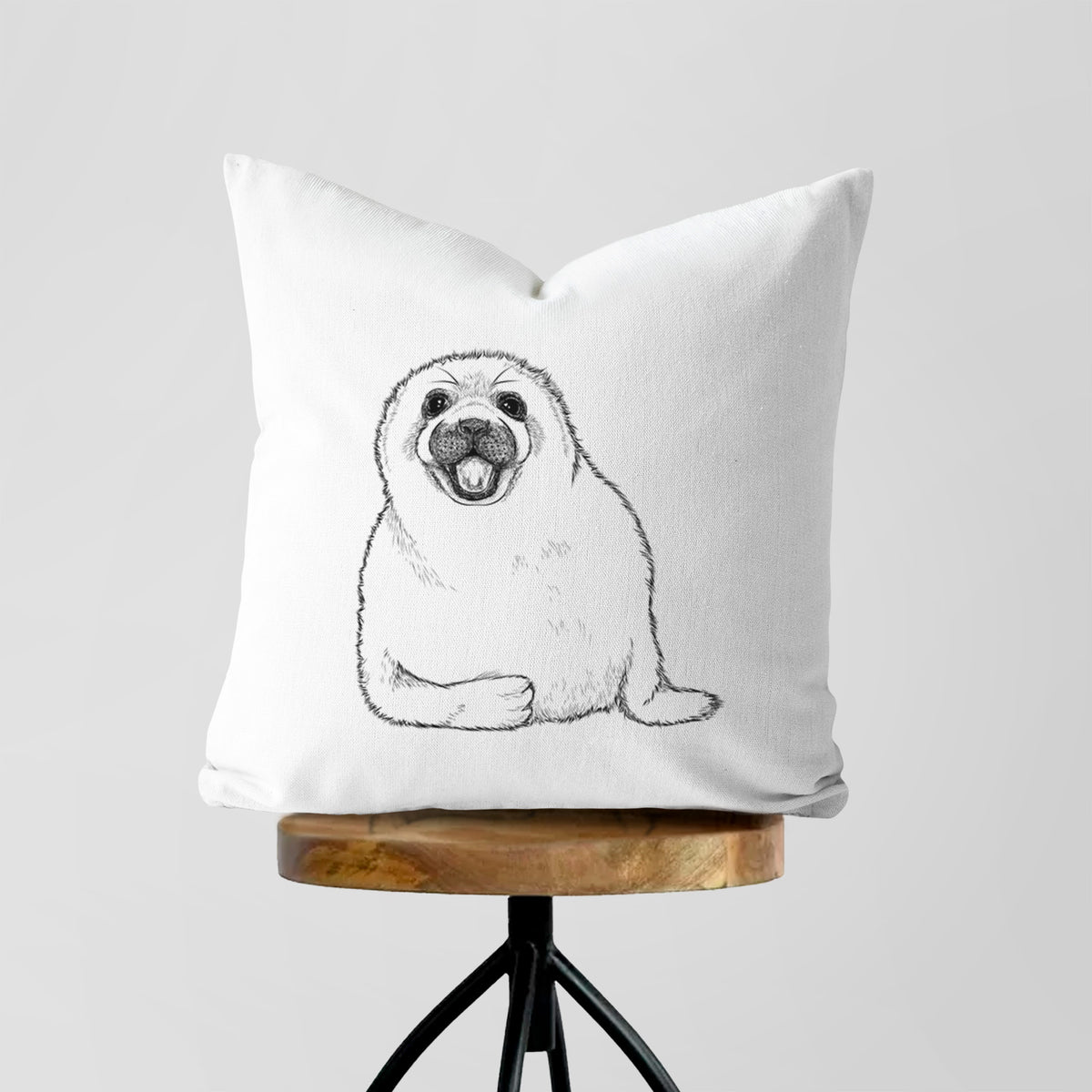 White pillow cover printed with a black ink illustration of a smiling harp seal pup lying down, on a wooden stool.